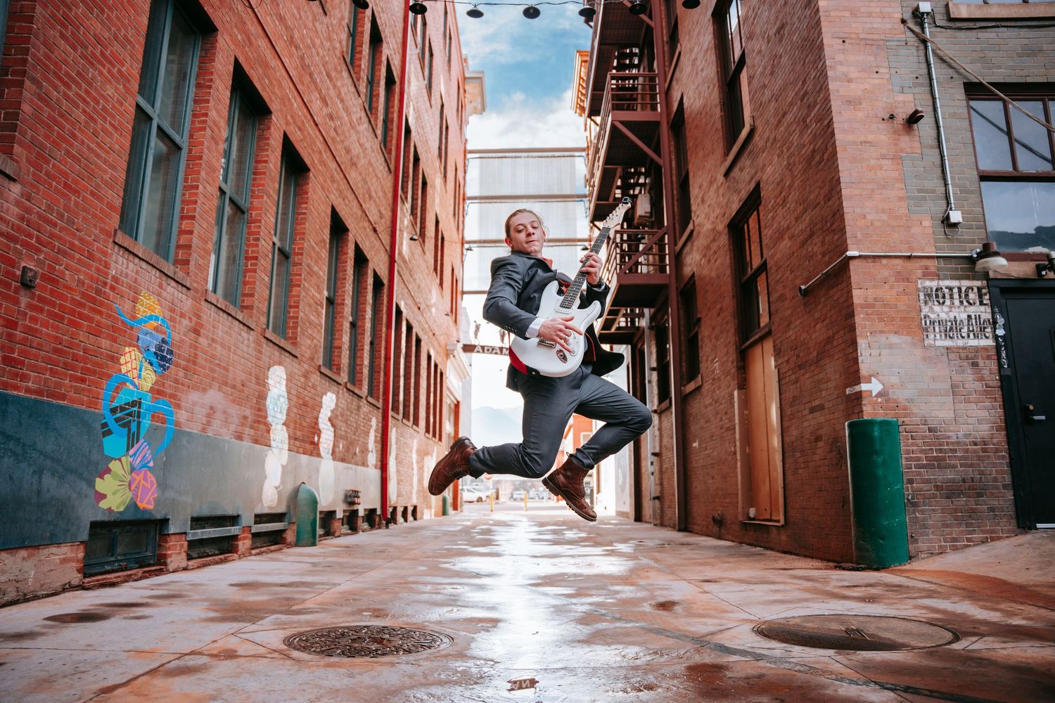Musician jumping joyfully in a wet urban alley holding a banjo, brick buildings and colorful street art in background.