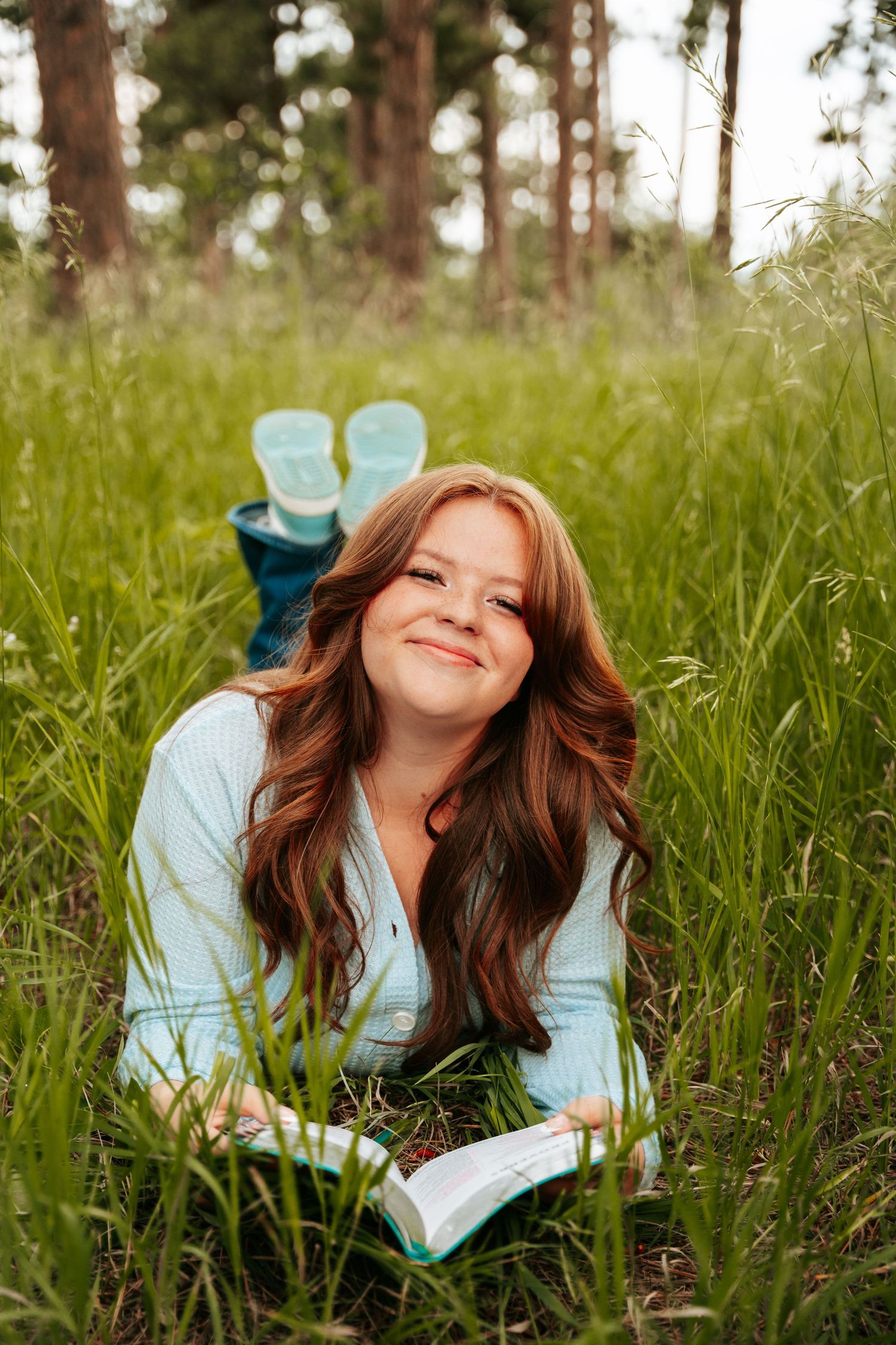 Smiling redhead woman lying in tall grass reading a book in forest meadow.
