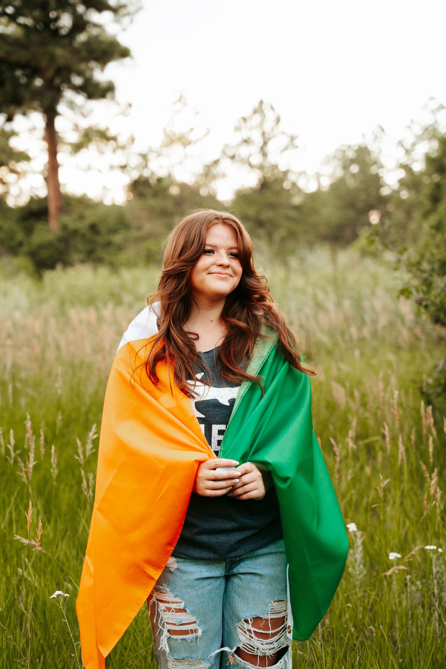 Smiling young woman with long brown hair wrapped in an Irish tricolor flag standing in a lush green meadow at sunset.