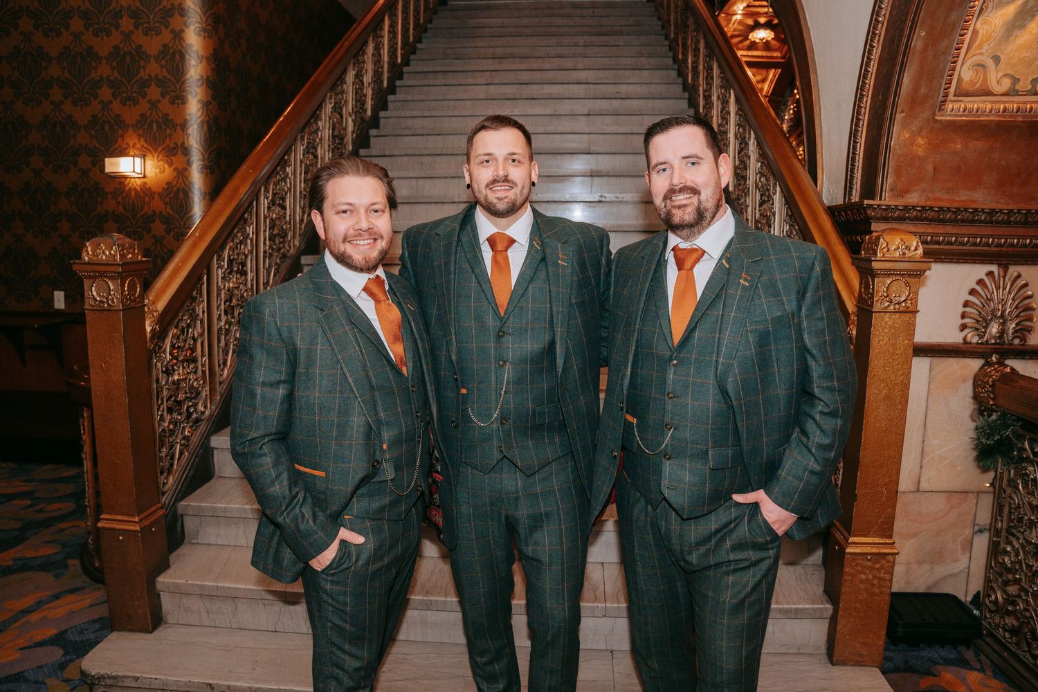 Three groomsmen in matching dark green plaid suits with orange ties on staircase.