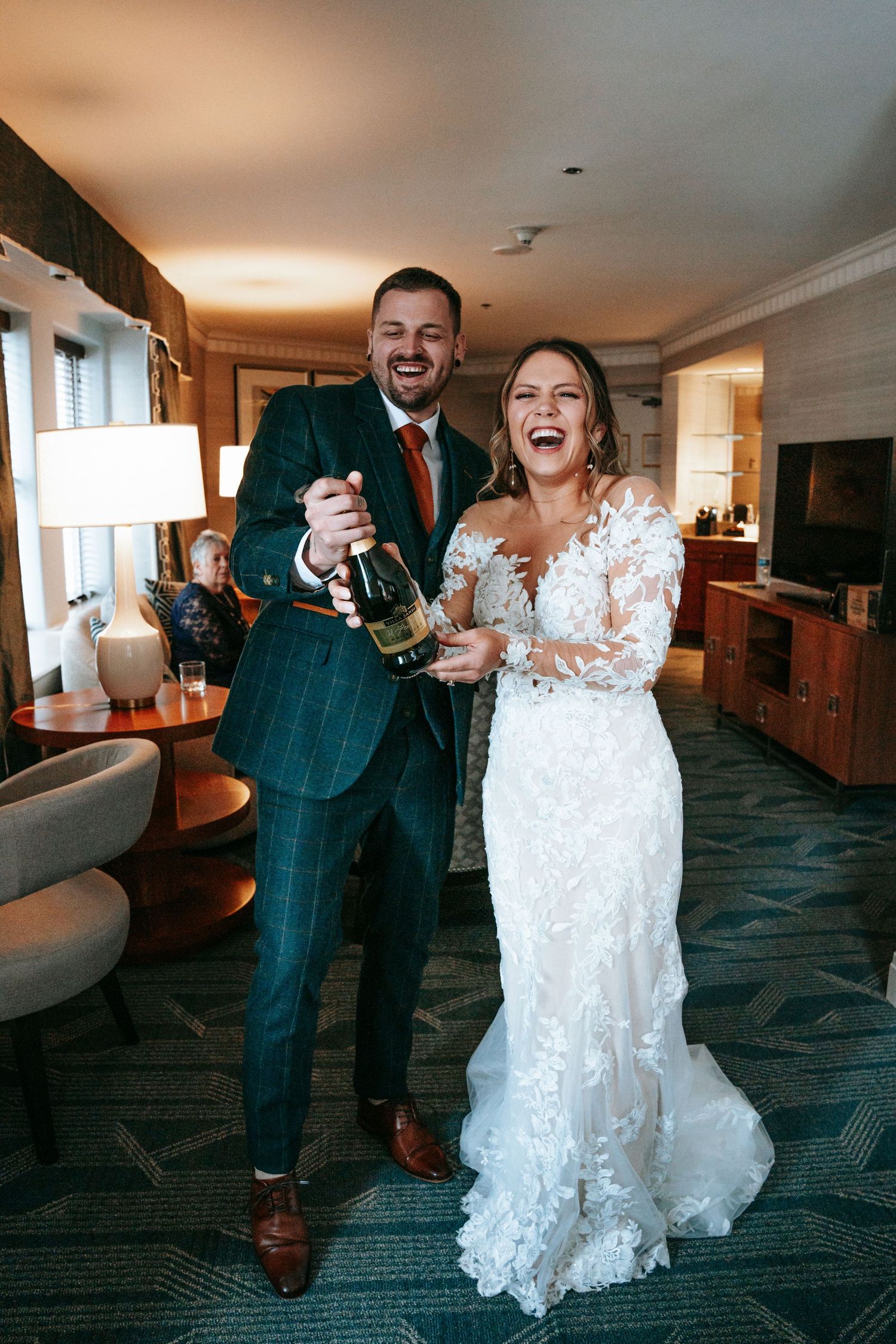A joyful bride and groom celebrating with a champagne bottle in a hotel suite.