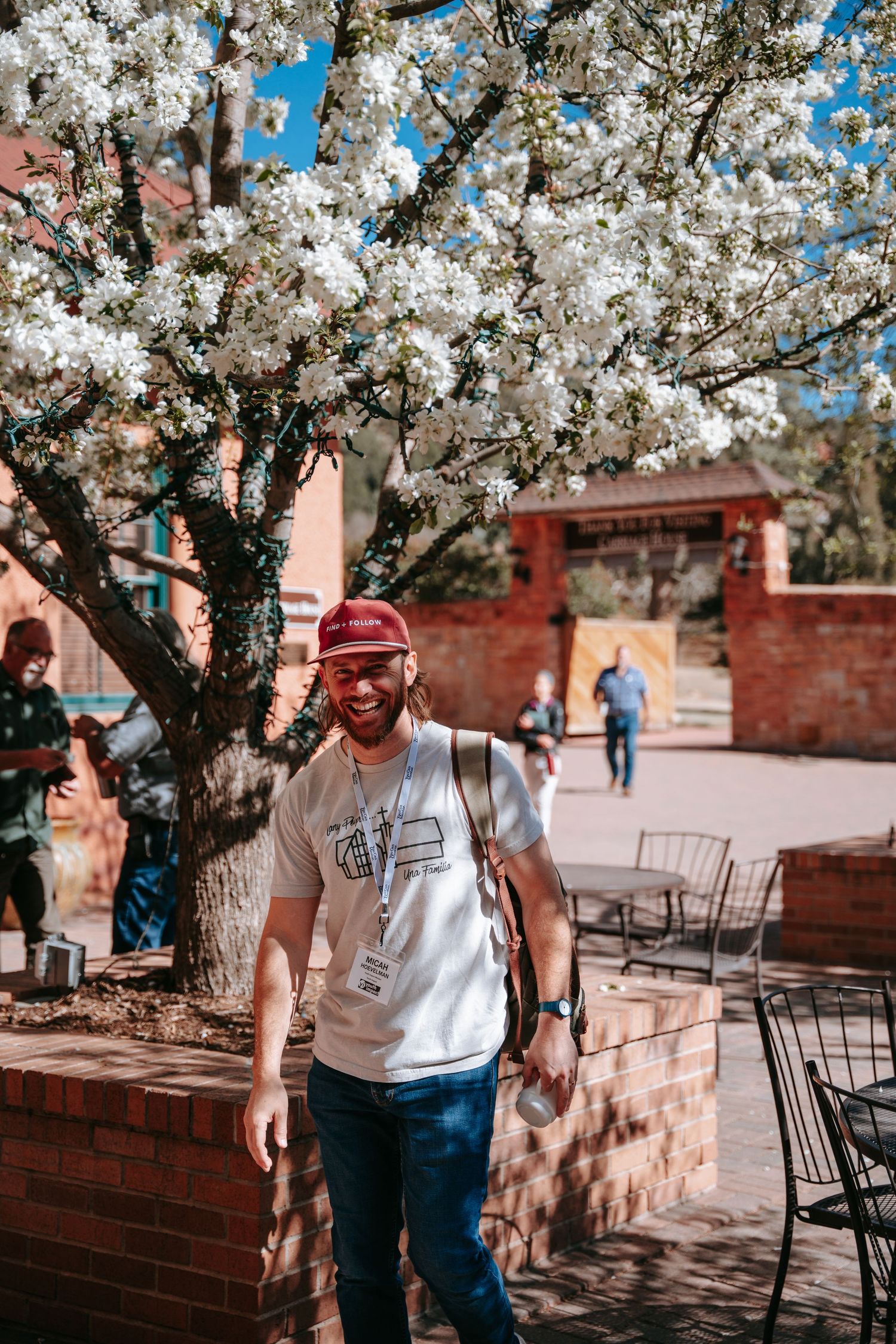 Smiling man in red cap and white tee walks near blooming white flowering tree on sunny day.