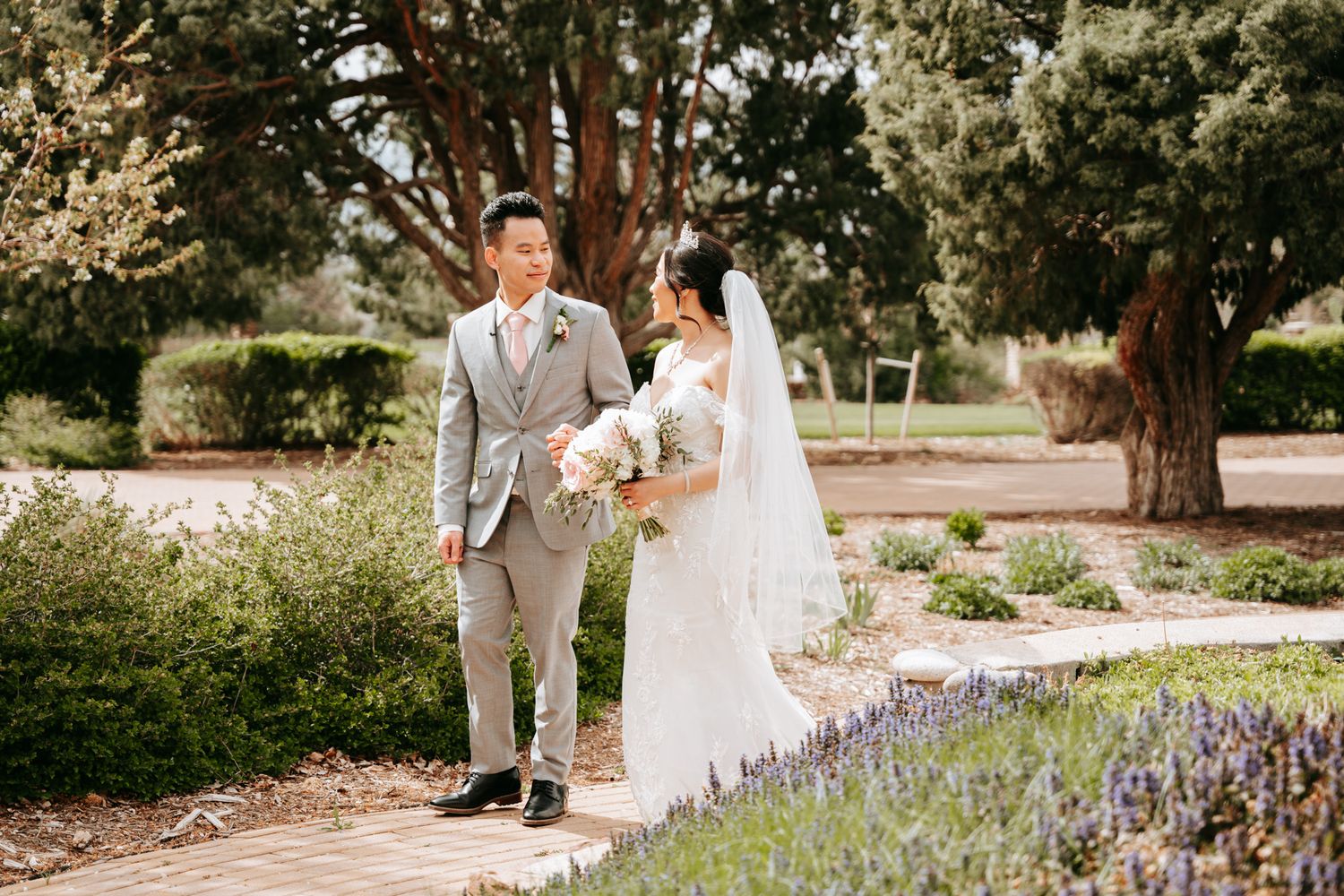 Asian wedding couple walking on garden path, groom in gray suit, bride in lace gown holding bouquet.
