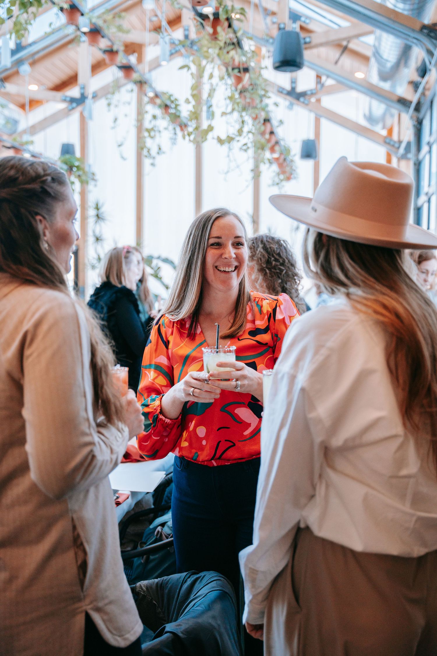 Smiling woman in red floral blouse holding drink at lively indoor social gathering with friends.