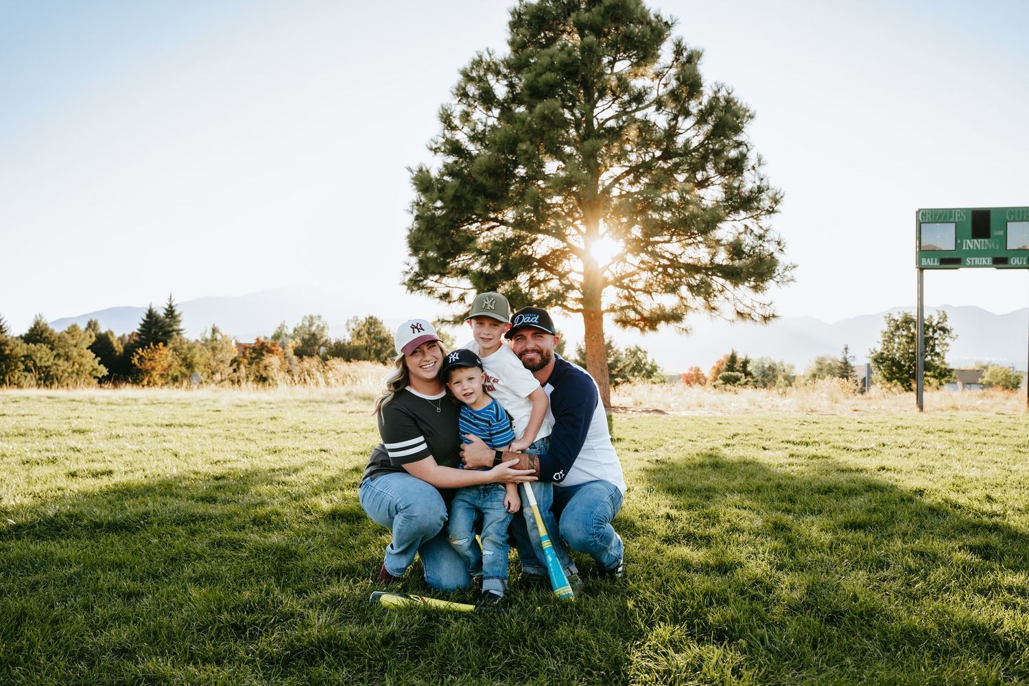 Family of four kneeling together on a sunny grassy baseball field, holding a bat with a tall pine tree and mountains in the background.