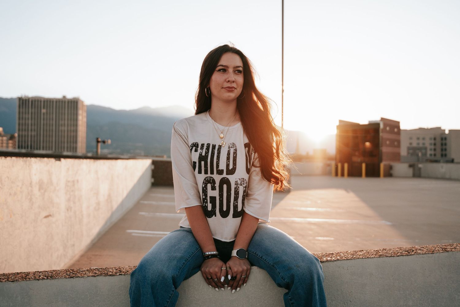 Young woman in Child of God tee sitting on rooftop ledge at sunset.