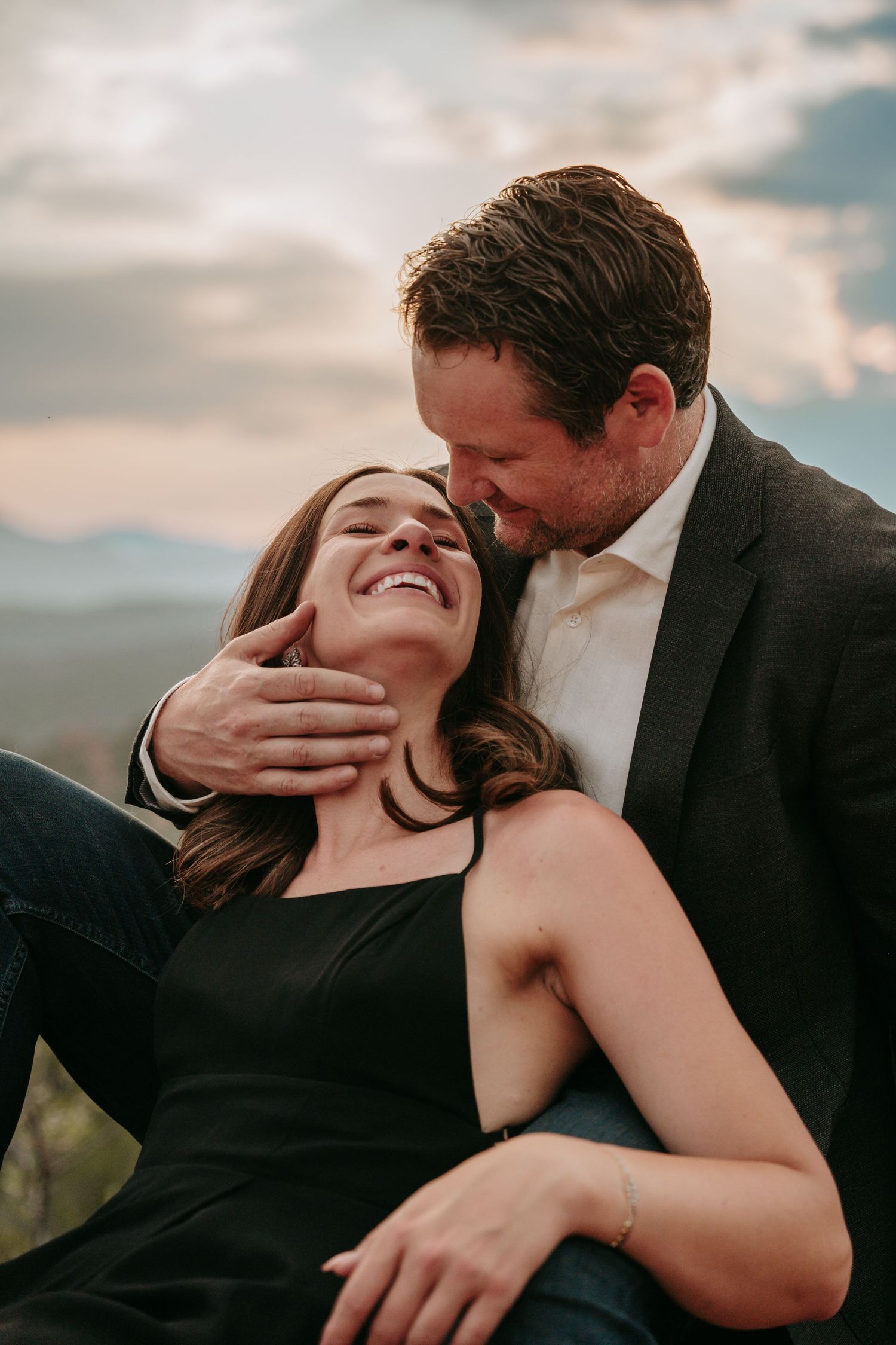 A couple shares a tender moment outdoors, woman laughing as man embraces her against a scenic mountain sunset backdrop.