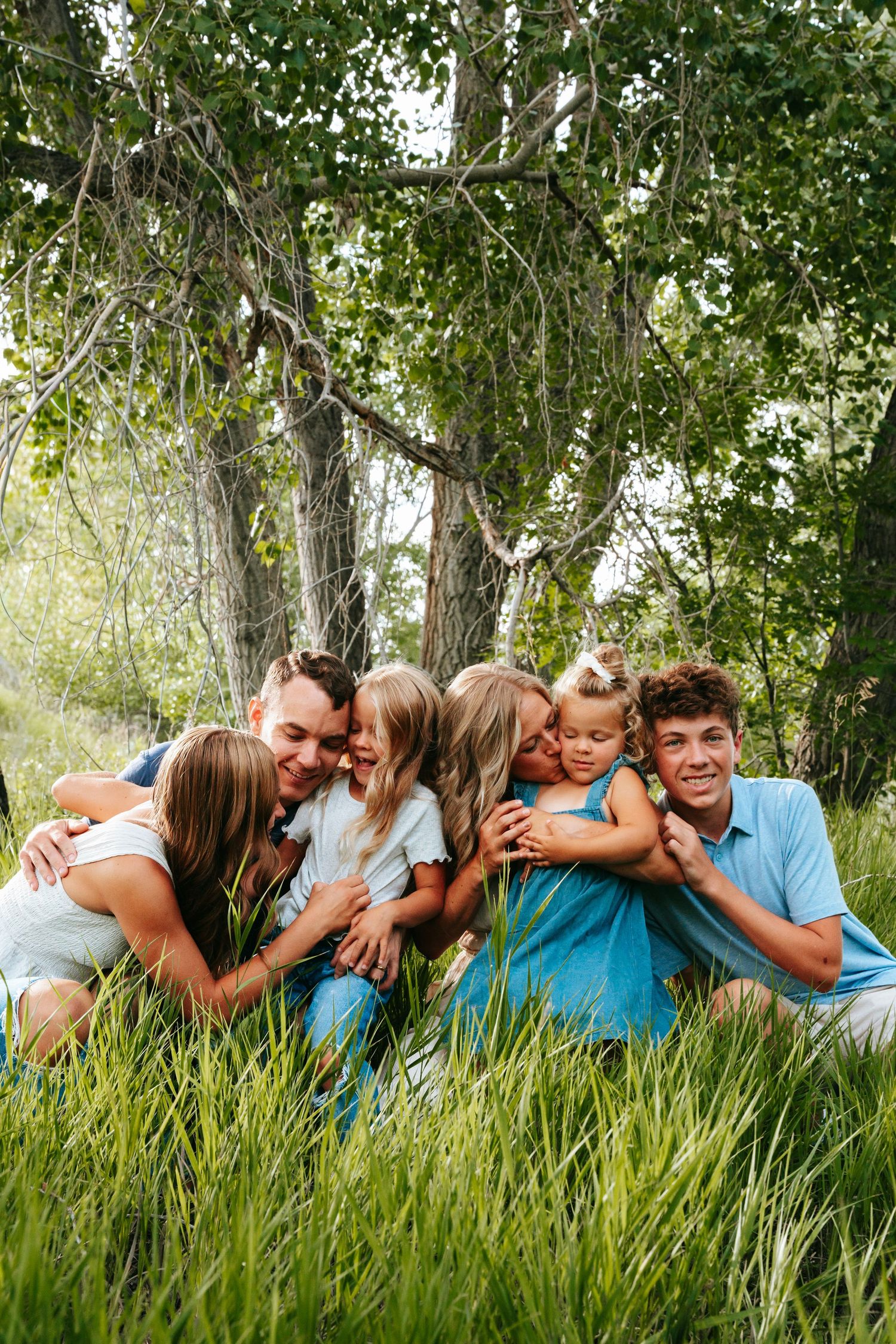 A joyful family sitting in tall green grass under trees, with children hugging parents and teens in a warm outdoor portrait.