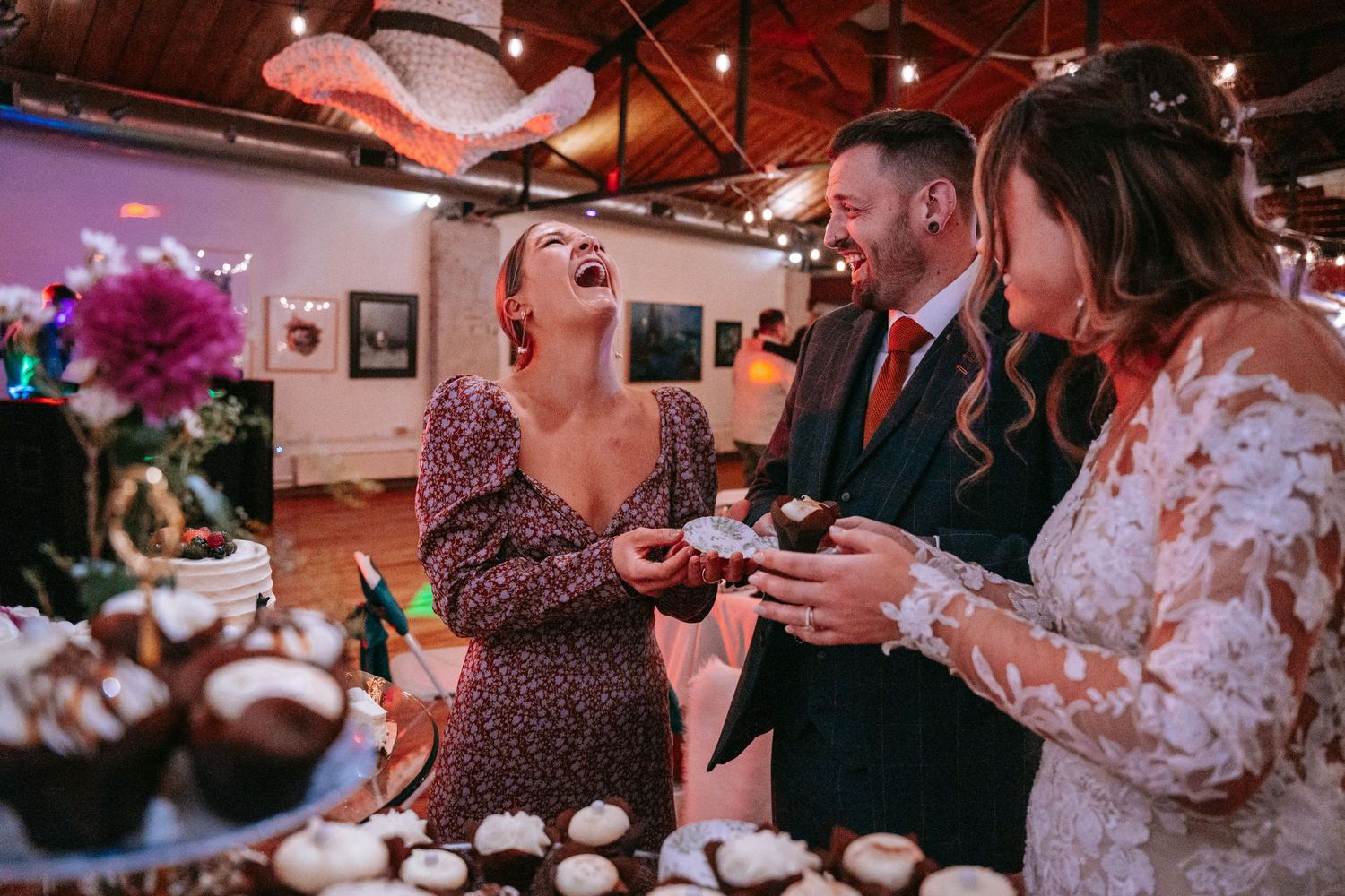 Guests laugh together near a cupcake dessert table at a lively wedding reception celebration.
