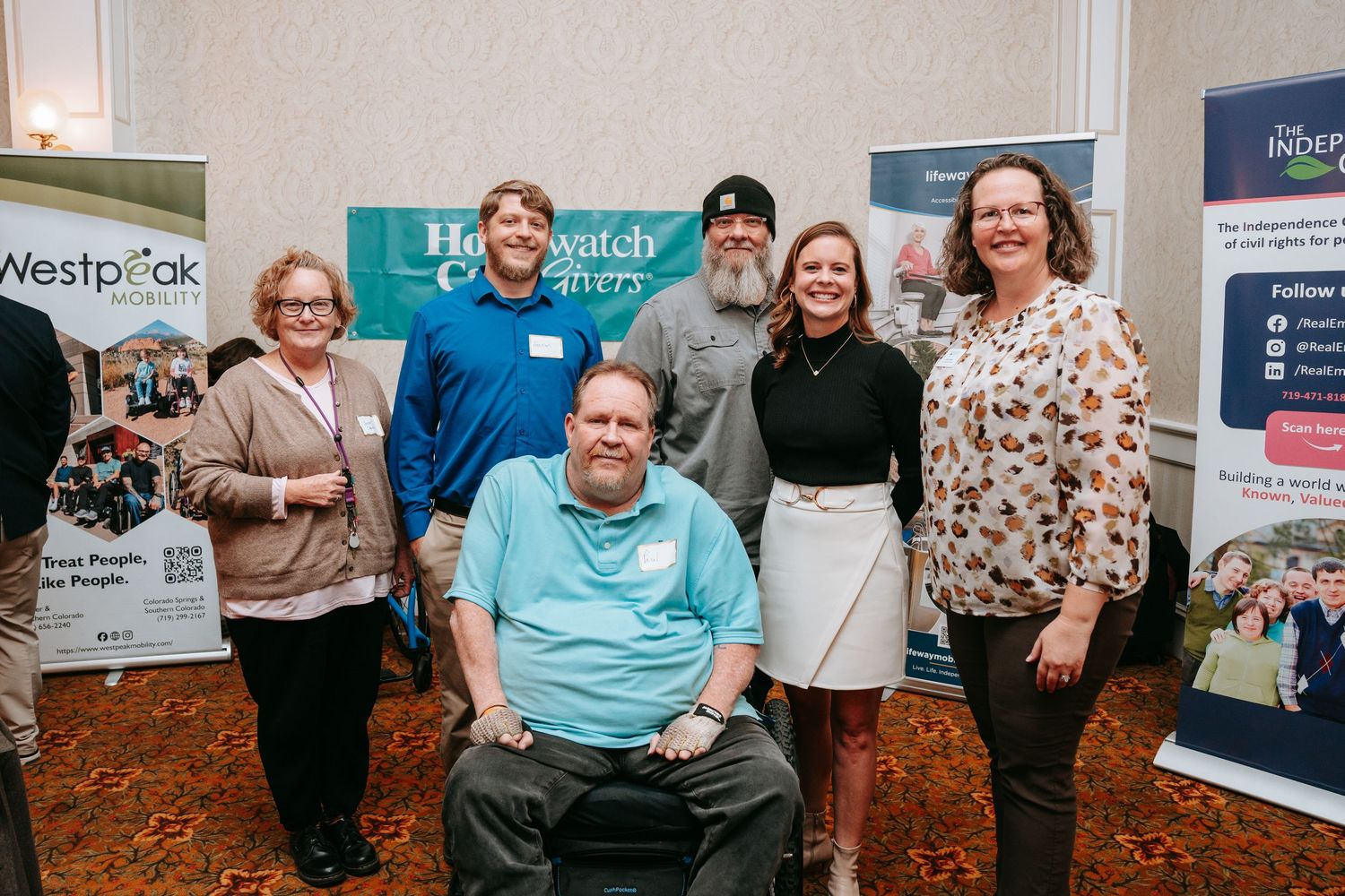 Six smiling people pose together at an indoor event, one seated in a wheelchair, with Westpeak and informational banners behind them.