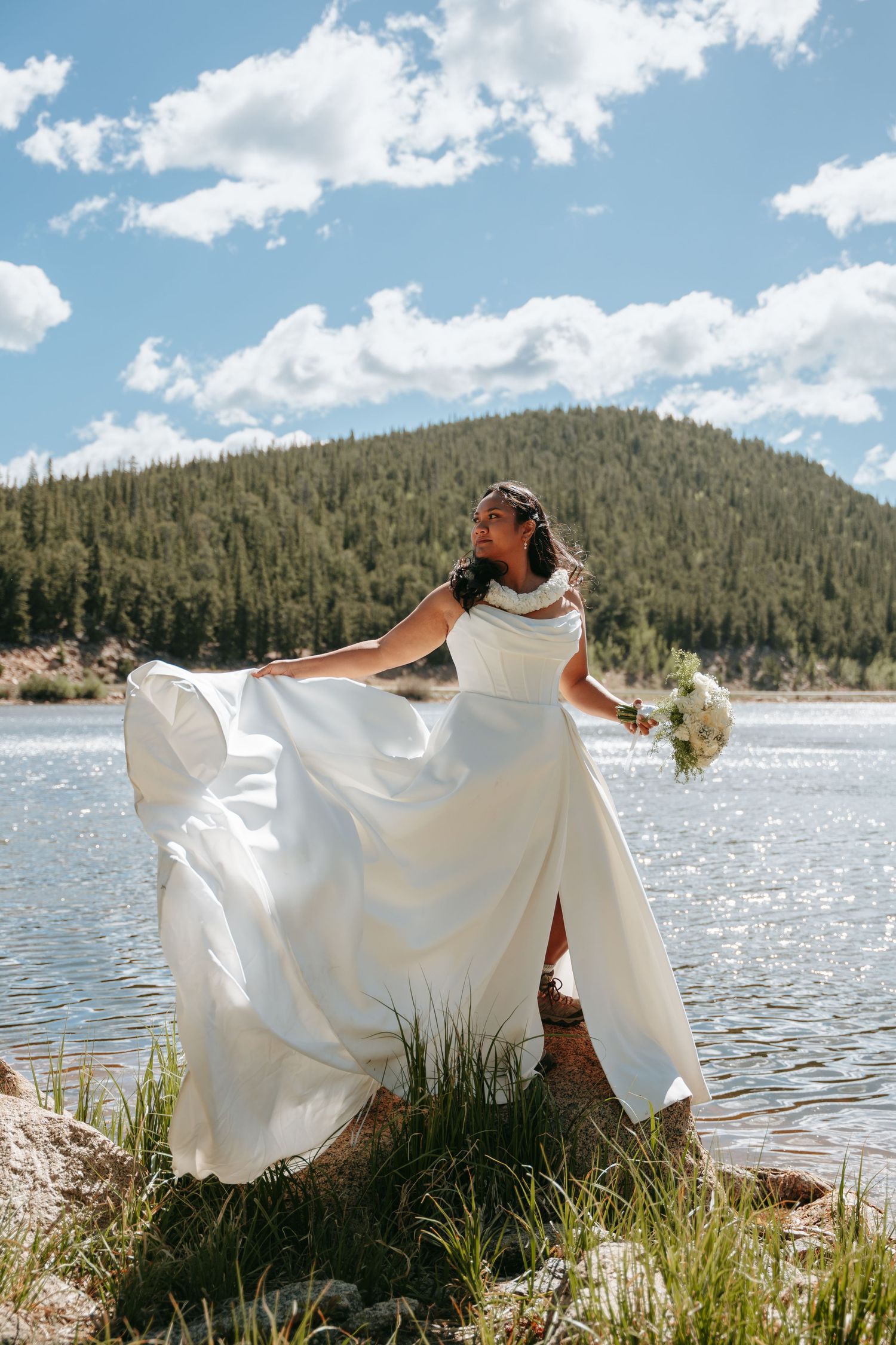 Bride in flowing white gown holding bouquet by mountain lake on sunny day.