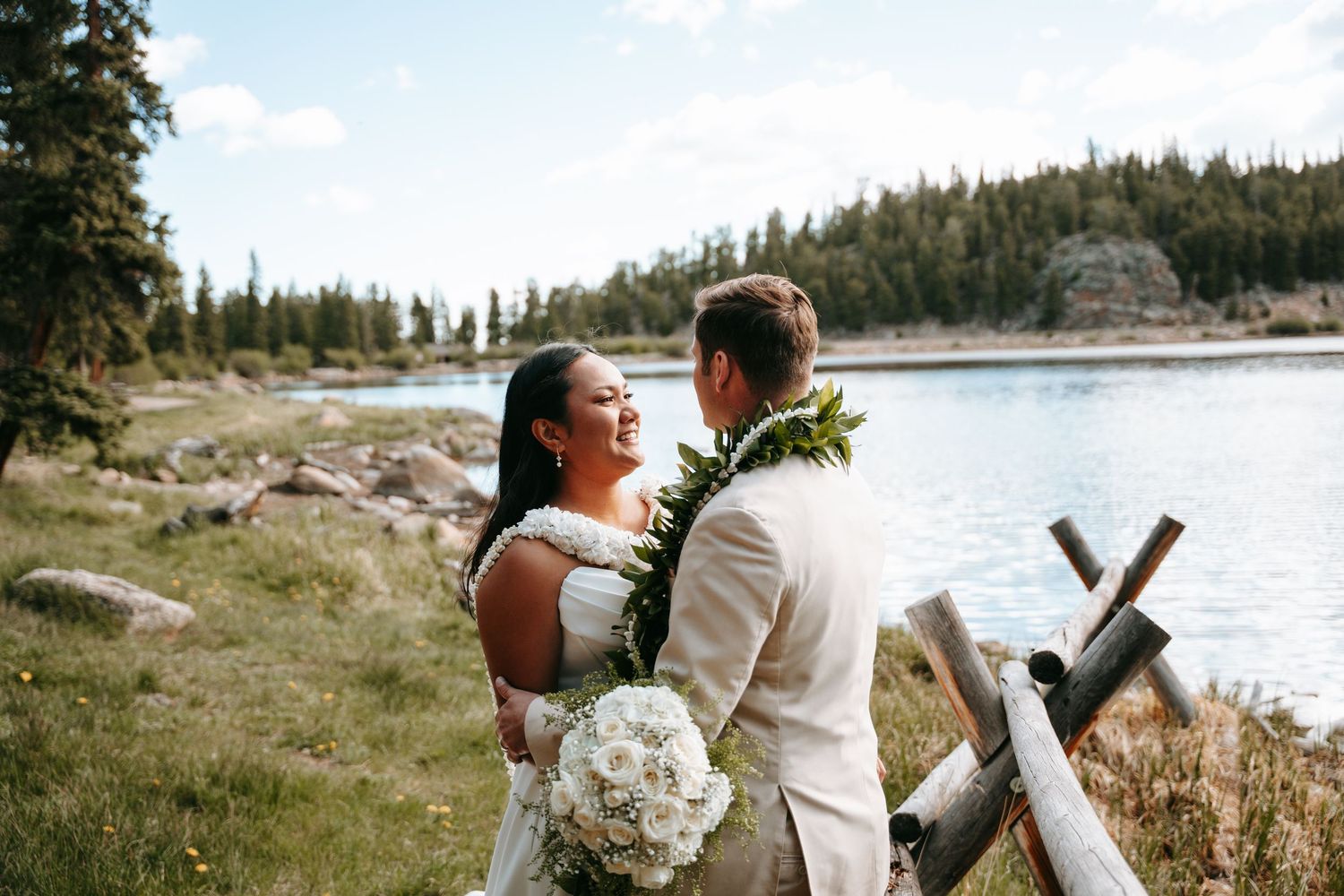 Bride and groom share a joyful moment by a mountain lake, bride holding white bouquet, groom wearing floral lei.