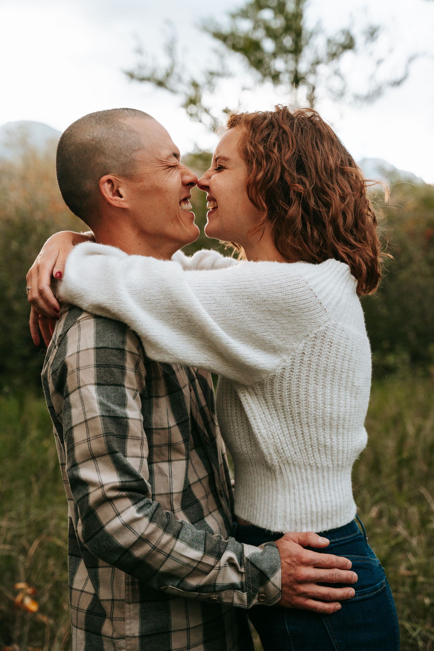 Couple embracing and touching noses in a lush green field with mountains behind.