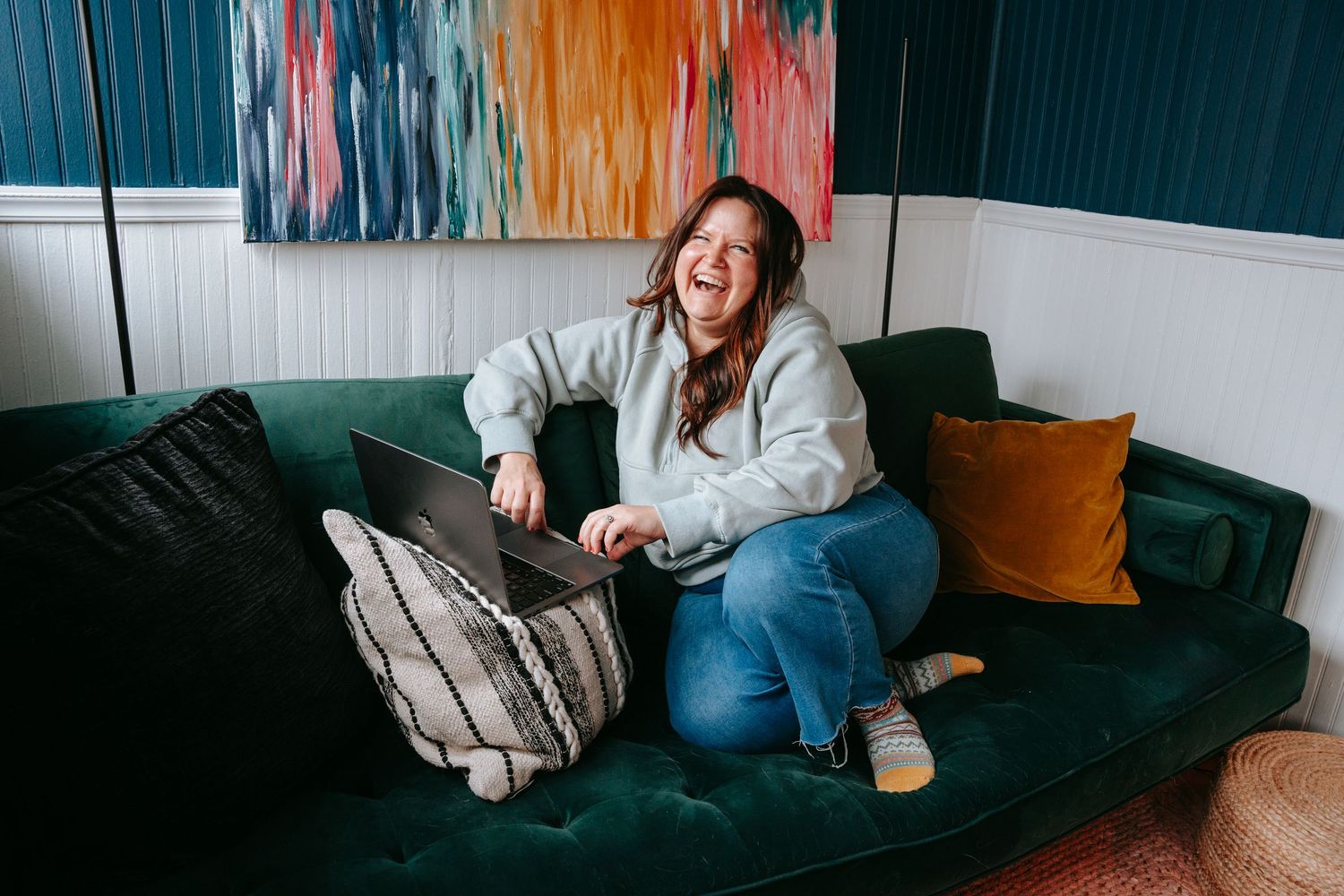 Joyful woman laughing with laptop on green velvet sofa with colorful abstract painting on wall behind.