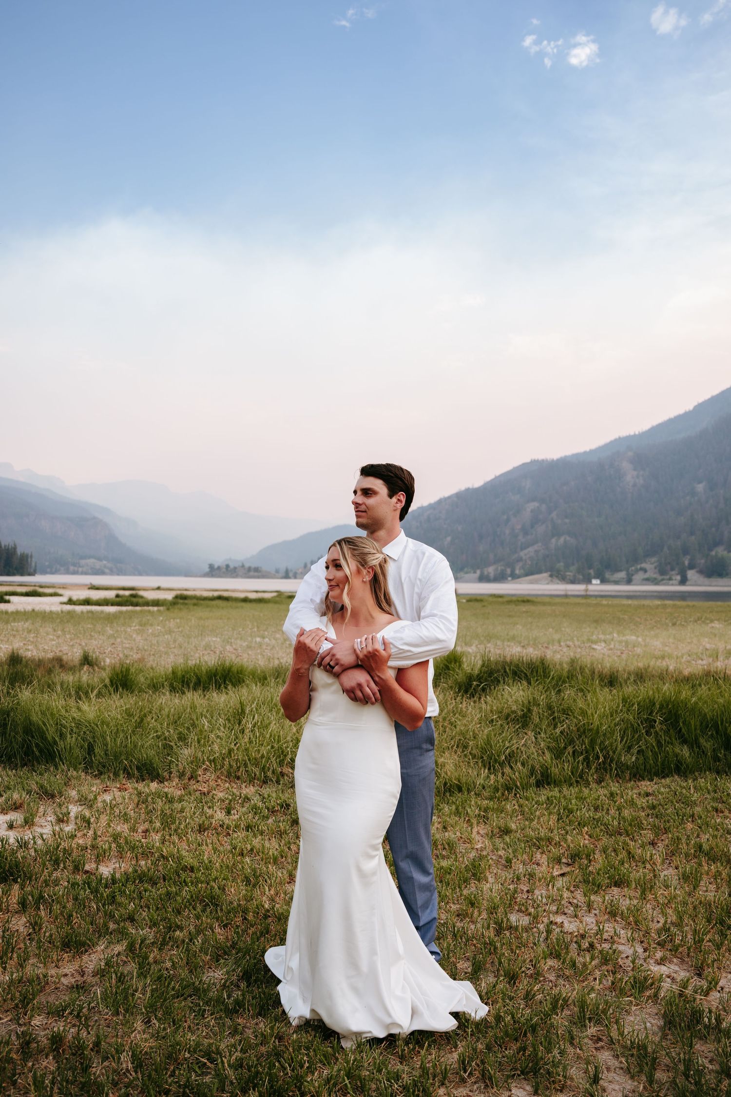 Bride and groom embracing in a mountain meadow at dusk, wearing wedding attire with scenic lake views.