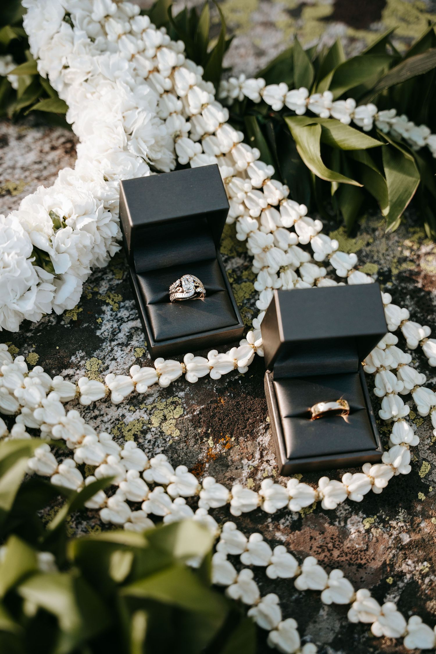 Two wedding rings in black boxes surrounded by white floral garlands and green leaves on a stone surface.