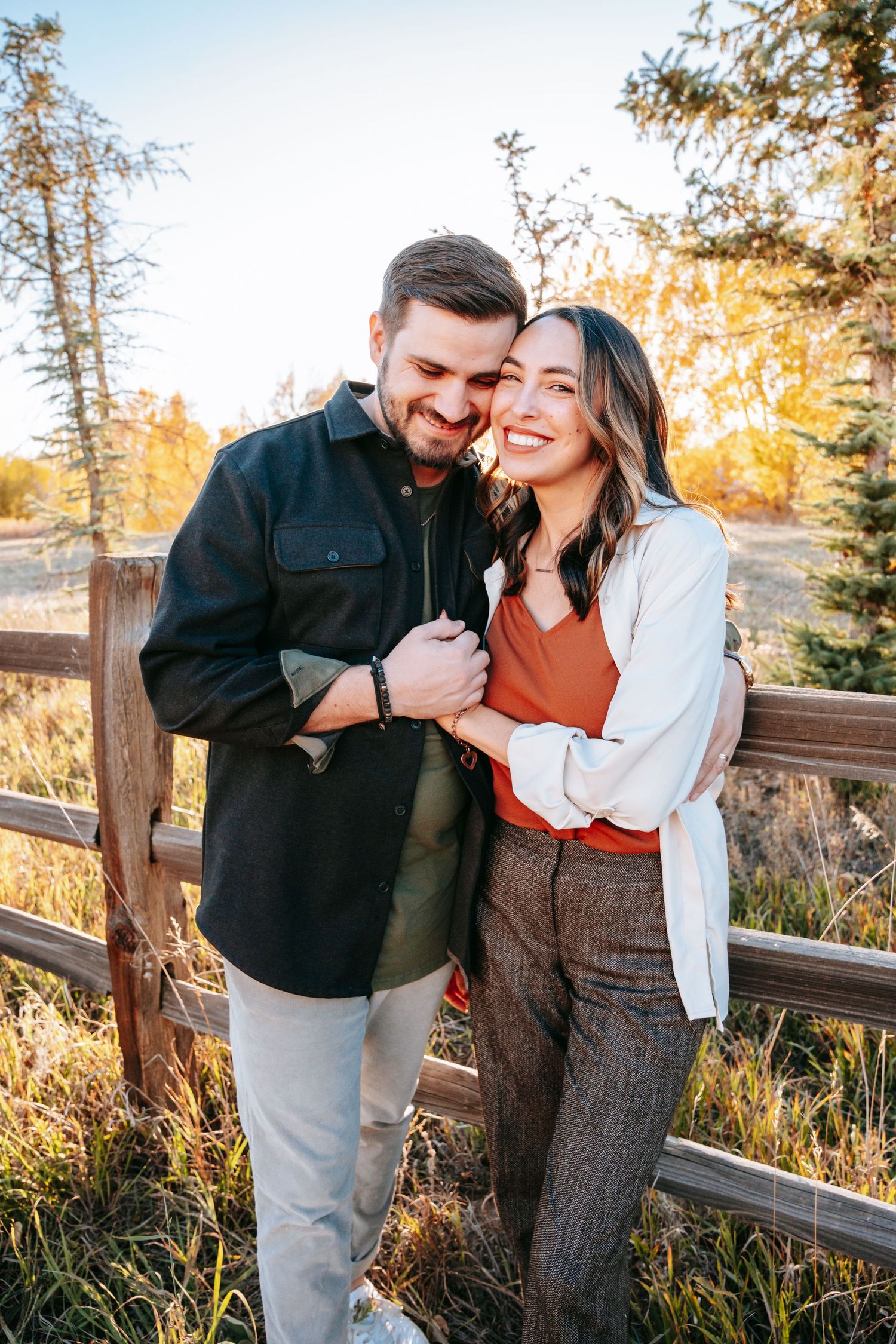 Happy couple embracing and laughing by a rustic wooden fence surrounded by autumn foliage.
