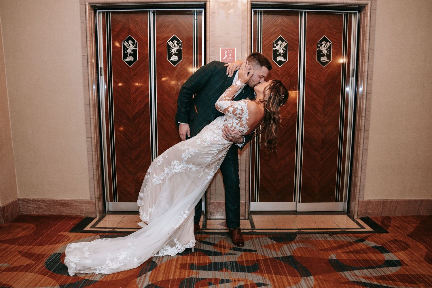 Bride and groom share a romantic dip kiss in front of elegant elevator doors at their wedding venue.