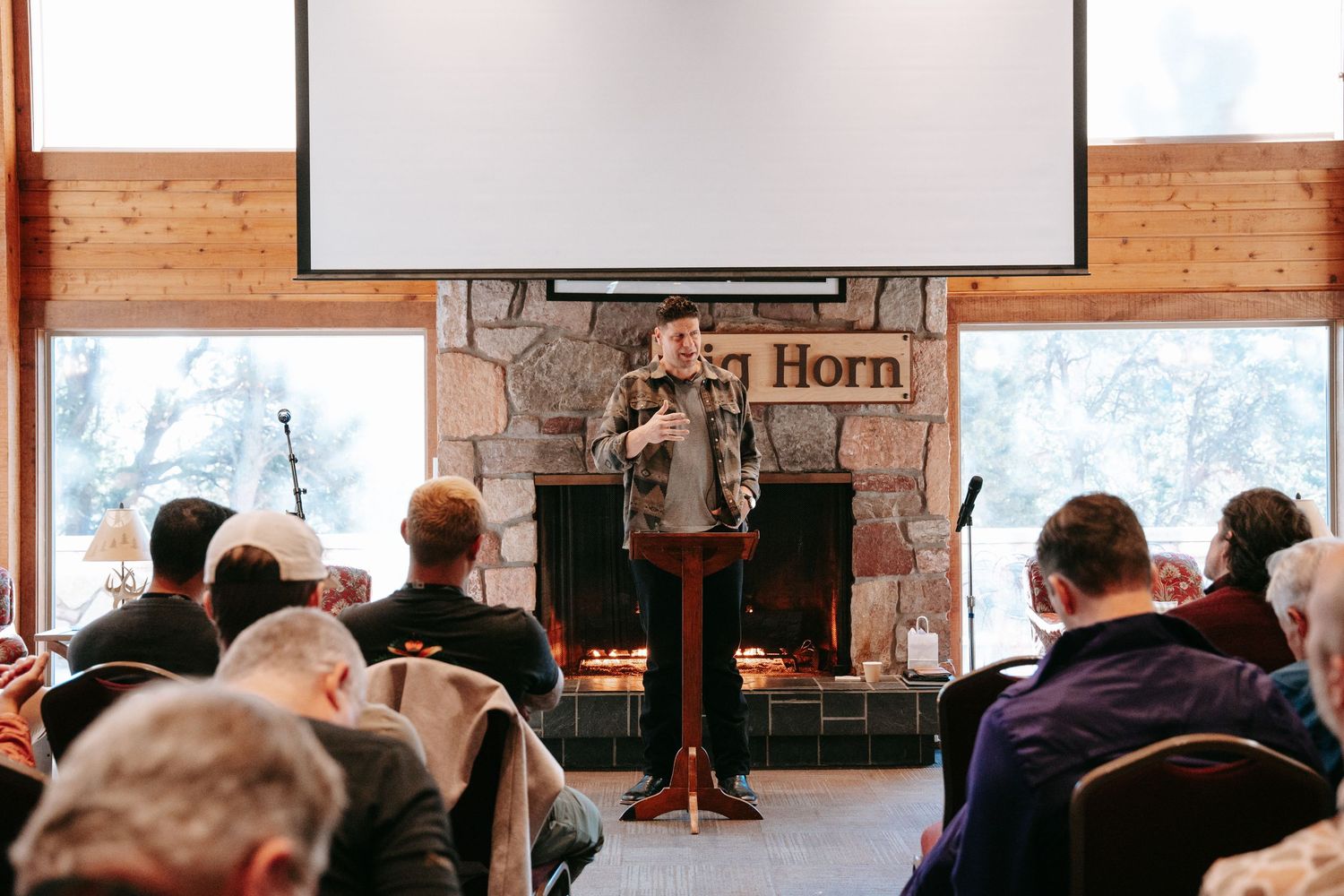 A speaker stands at a wooden podium in a lodge-style room with a stone fireplace while an audience watches from seated rows.