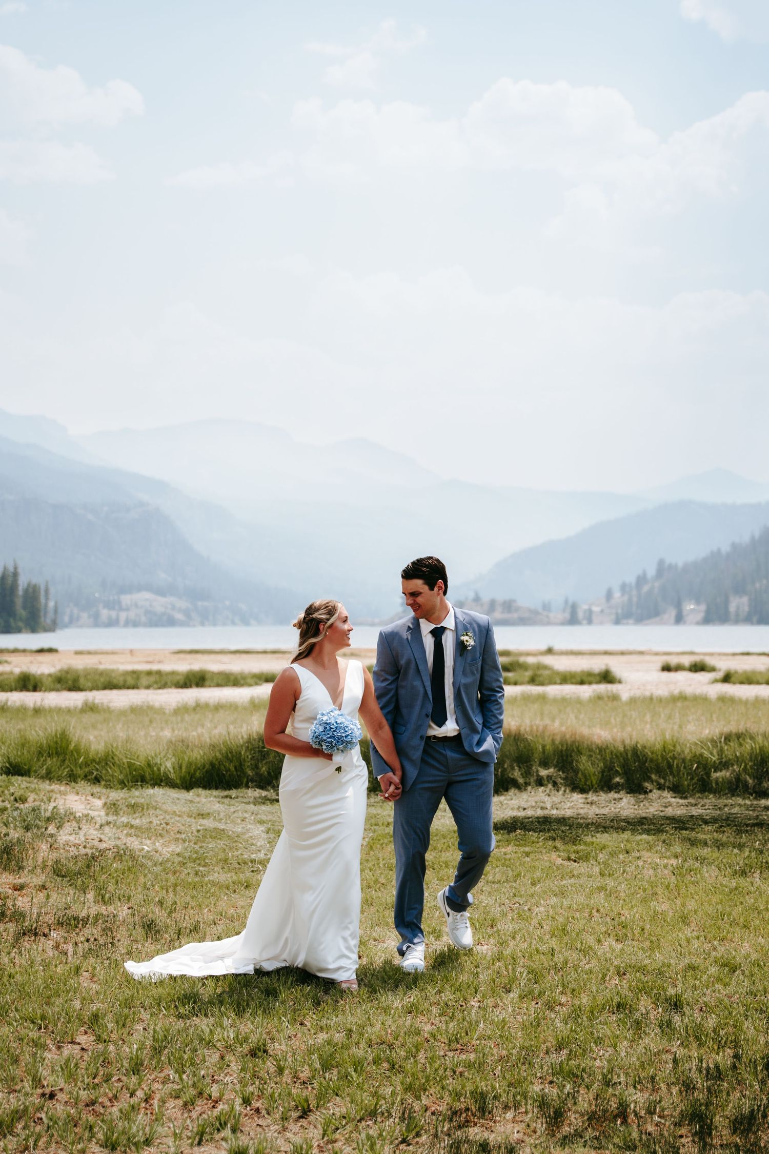 Bride and groom holding hands in a mountain meadow, bride holding blue hydrangeas bouquet with lake and mountains behind.
