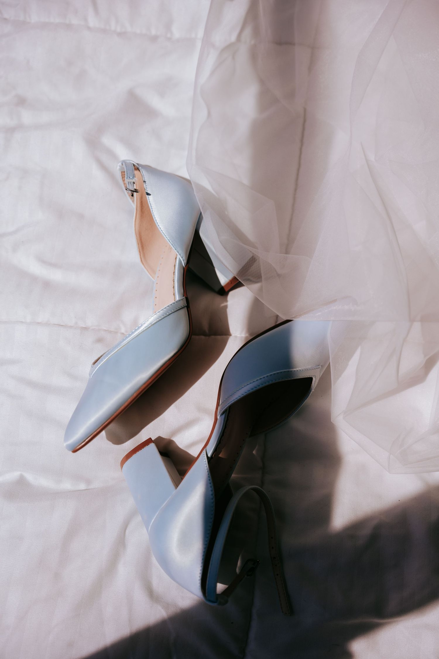 Light blue satin block heel wedding shoes with ankle strap resting on white bridal veil fabric.