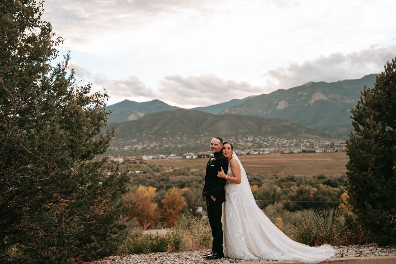 Bride and groom pose together among autumn trees with scenic mountain landscape in the background.