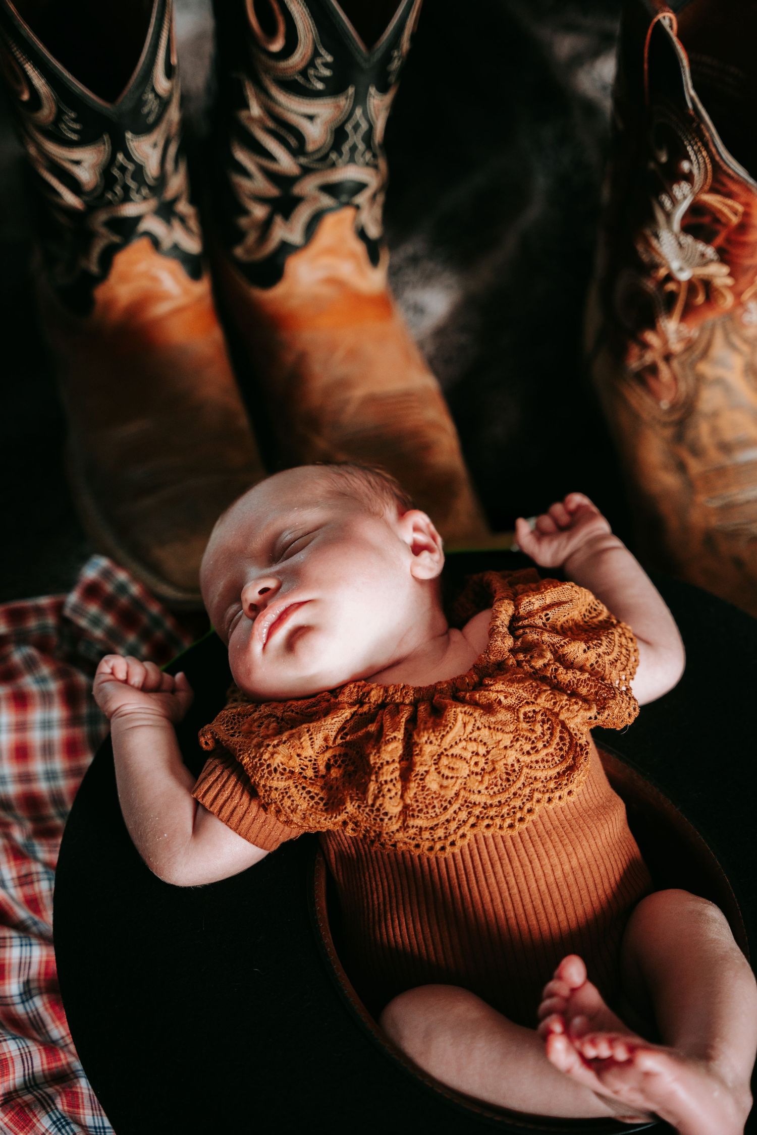 Sleeping newborn baby in rust orange lace onesie resting between two pairs of cowboy boots.