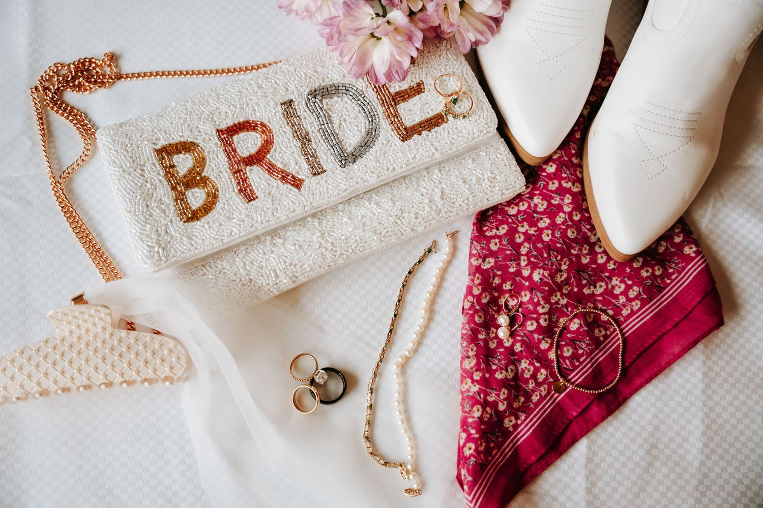 Flat lay of bridal accessories including BRIDE clutch, white boots, rings, and floral bandana.