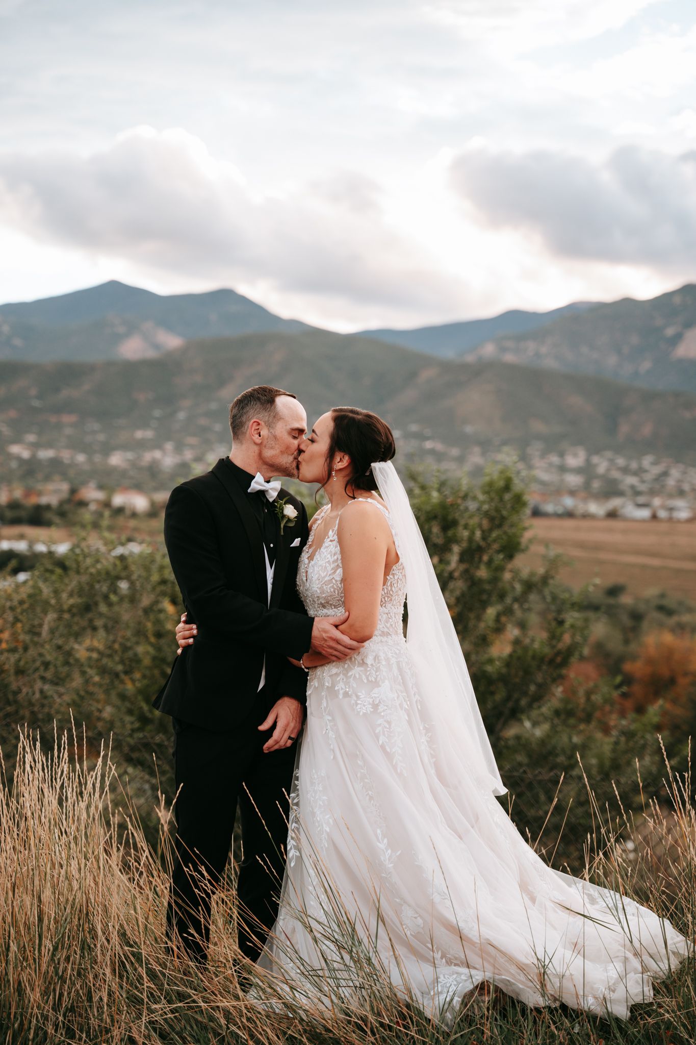 Bride and groom share a kiss on a hilltop with stunning mountain and valley views behind them.