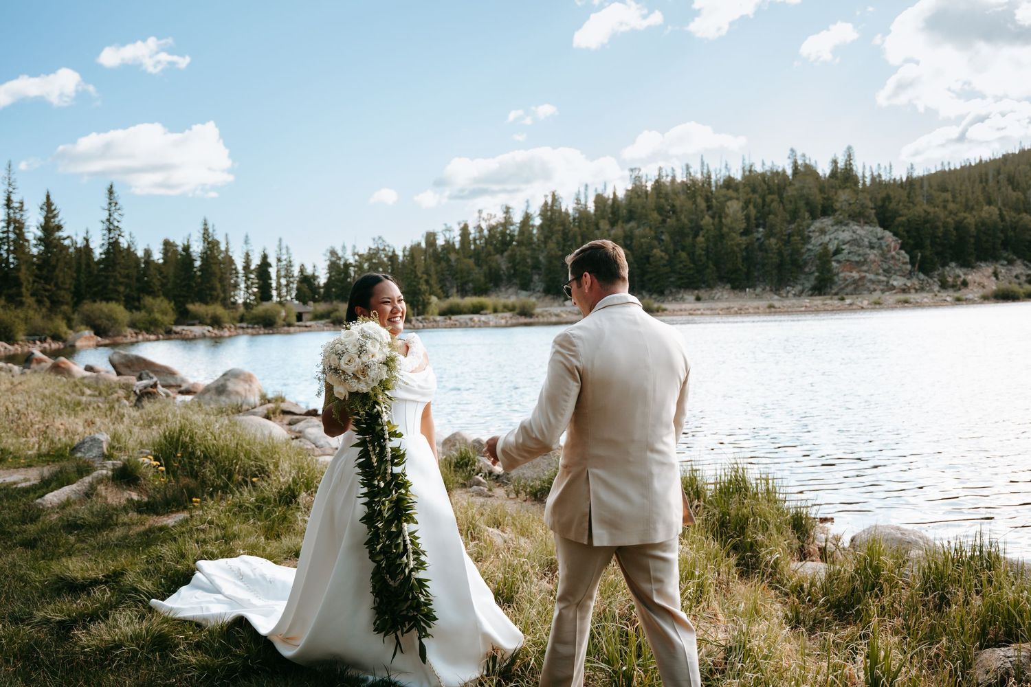 Bride and groom walk hand in hand beside a mountain lake surrounded by pine trees on their wedding day.