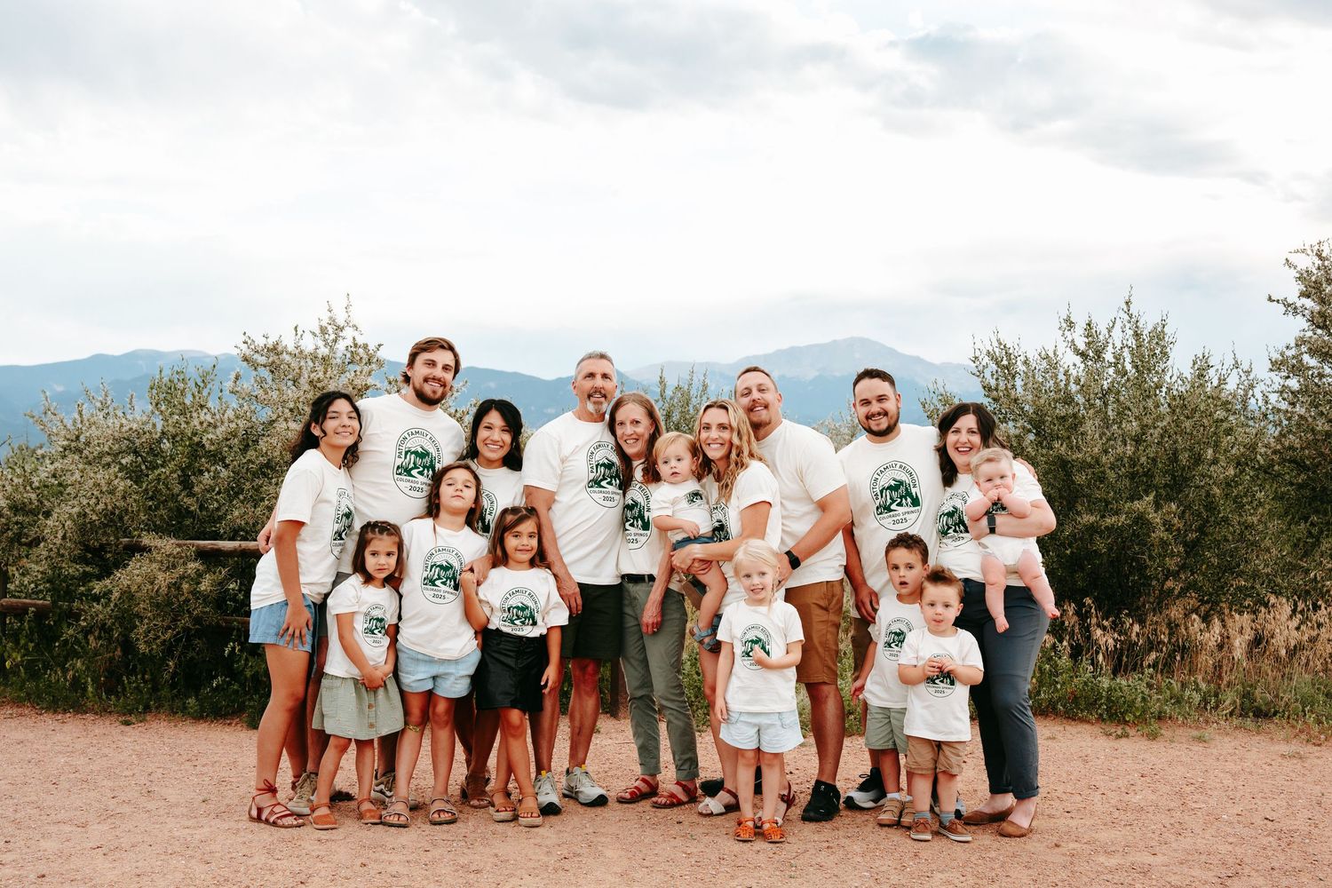 Large family group wearing matching white tree logo shirts posing together outdoors with mountain views in background.