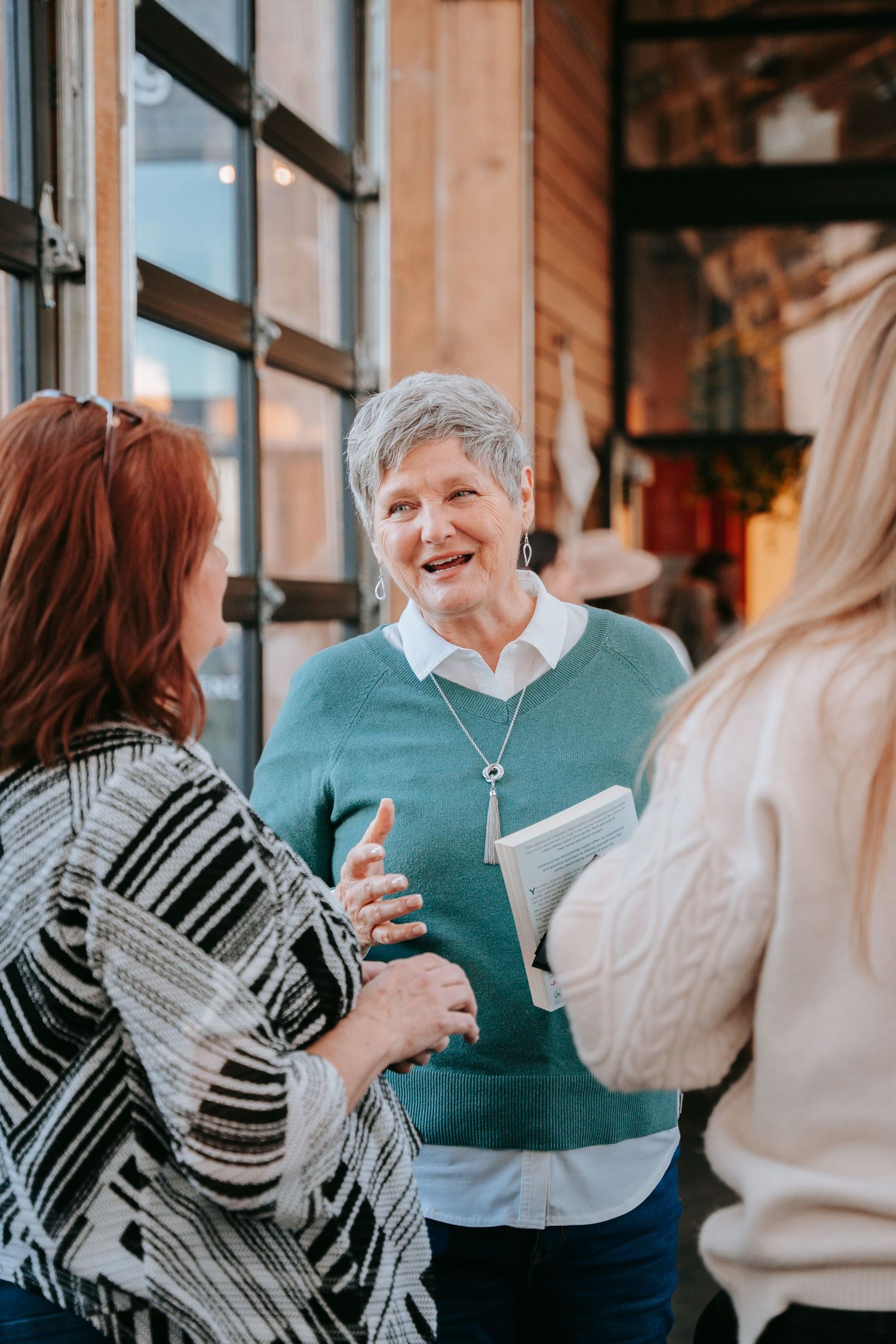 Three women conversing indoors, center woman in teal sweater holding a book and smiling warmly.