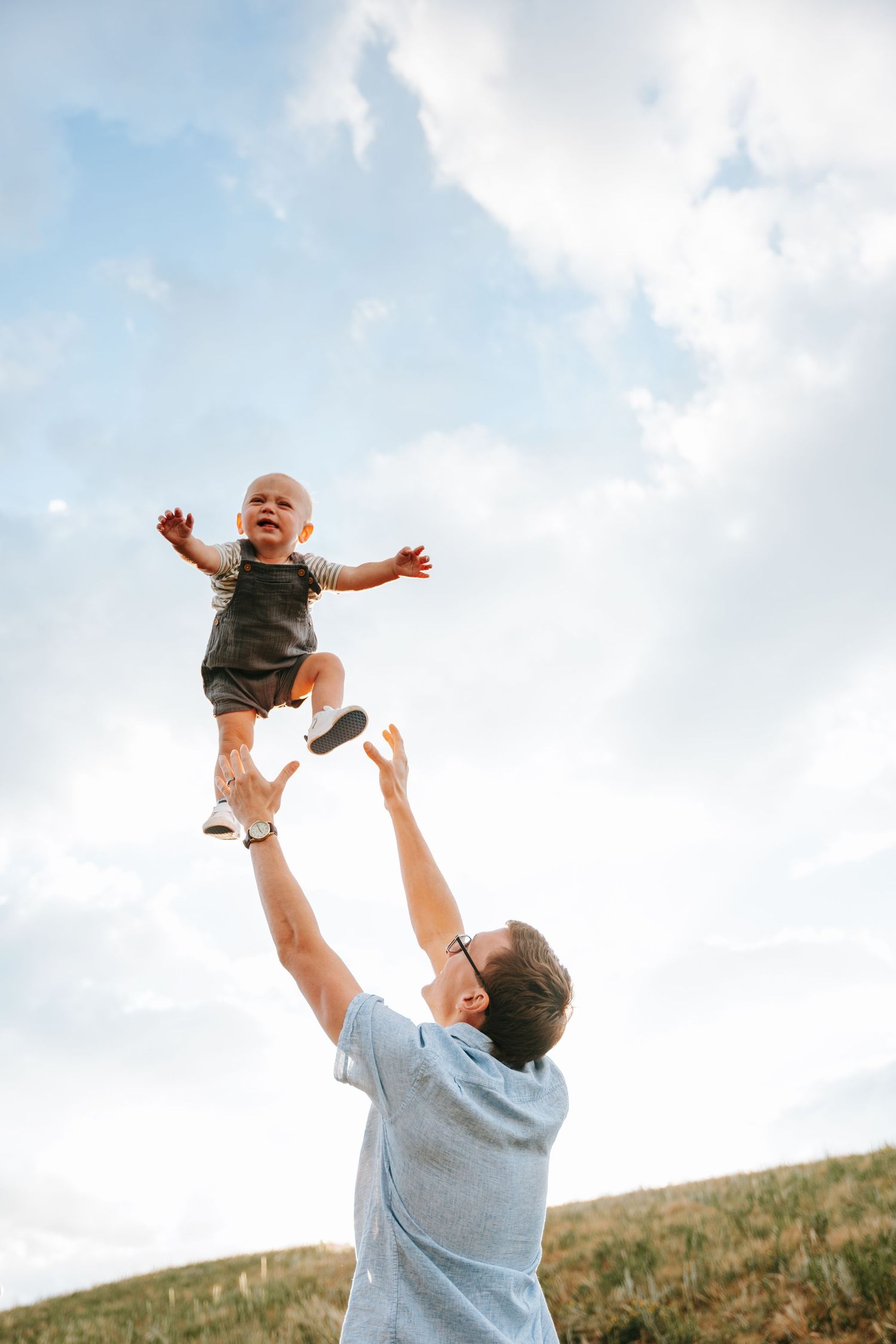 Father tossing laughing baby into the air against a bright cloudy sky in an open grassy field.
