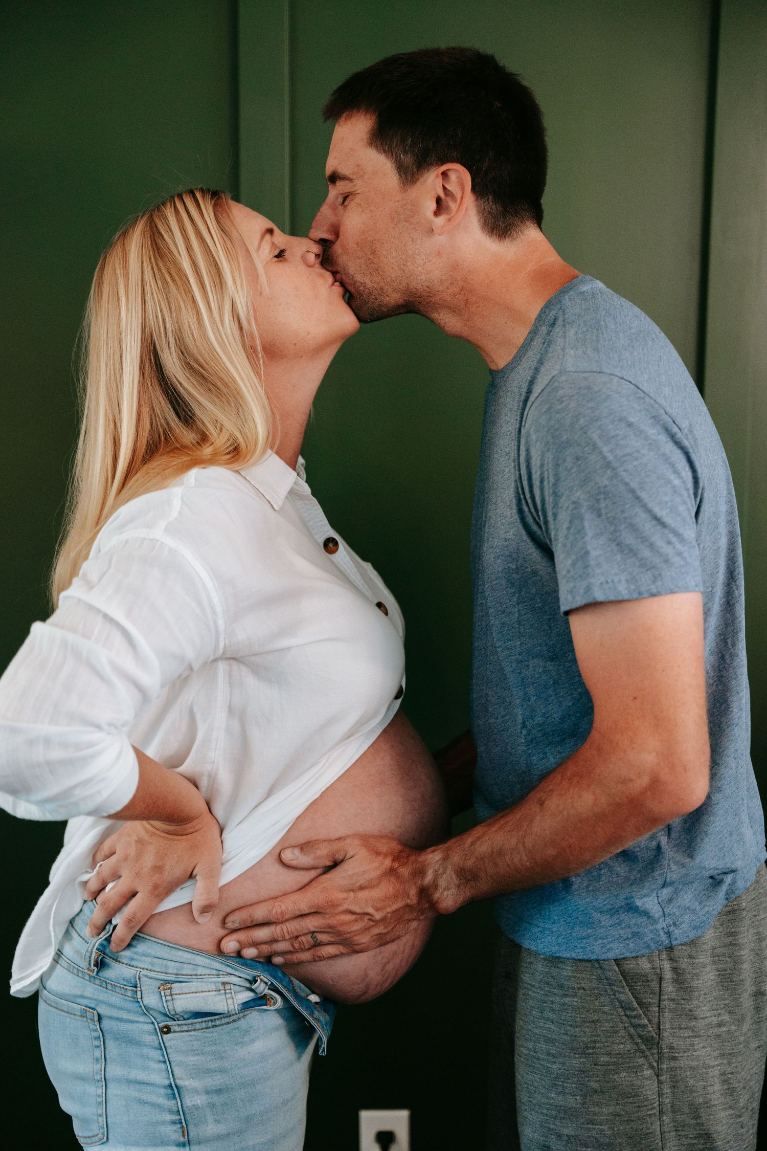 Pregnant couple kissing while cradling baby bump against green wall in maternity photo.