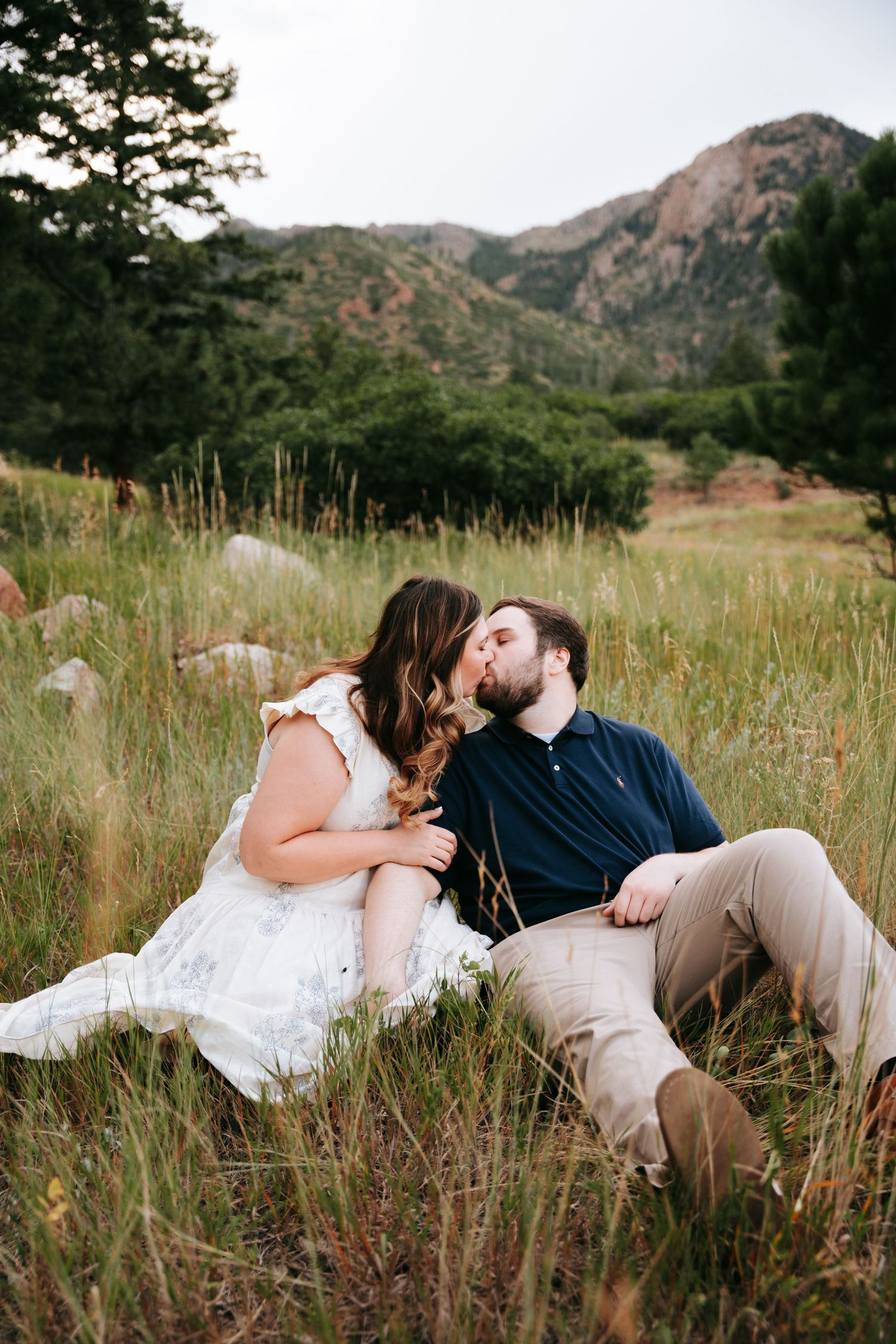 Couple kissing romantically while sitting in tall green grass with rocky mountain backdrop.