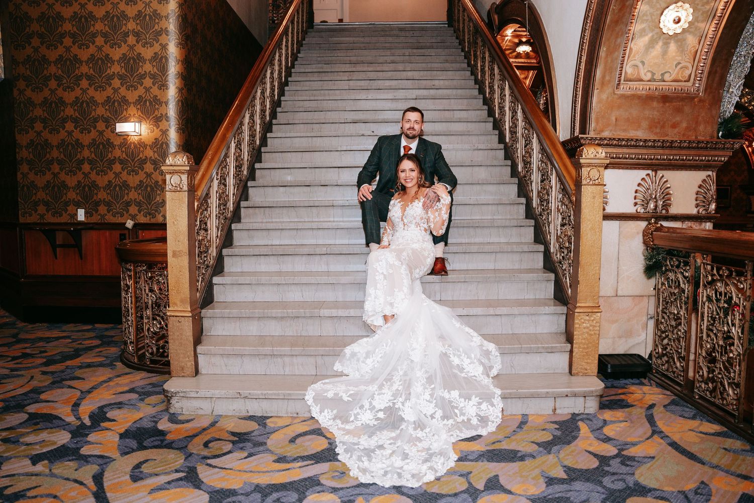 Bride in lace mermaid gown with long train poses with groom on ornate grand staircase.