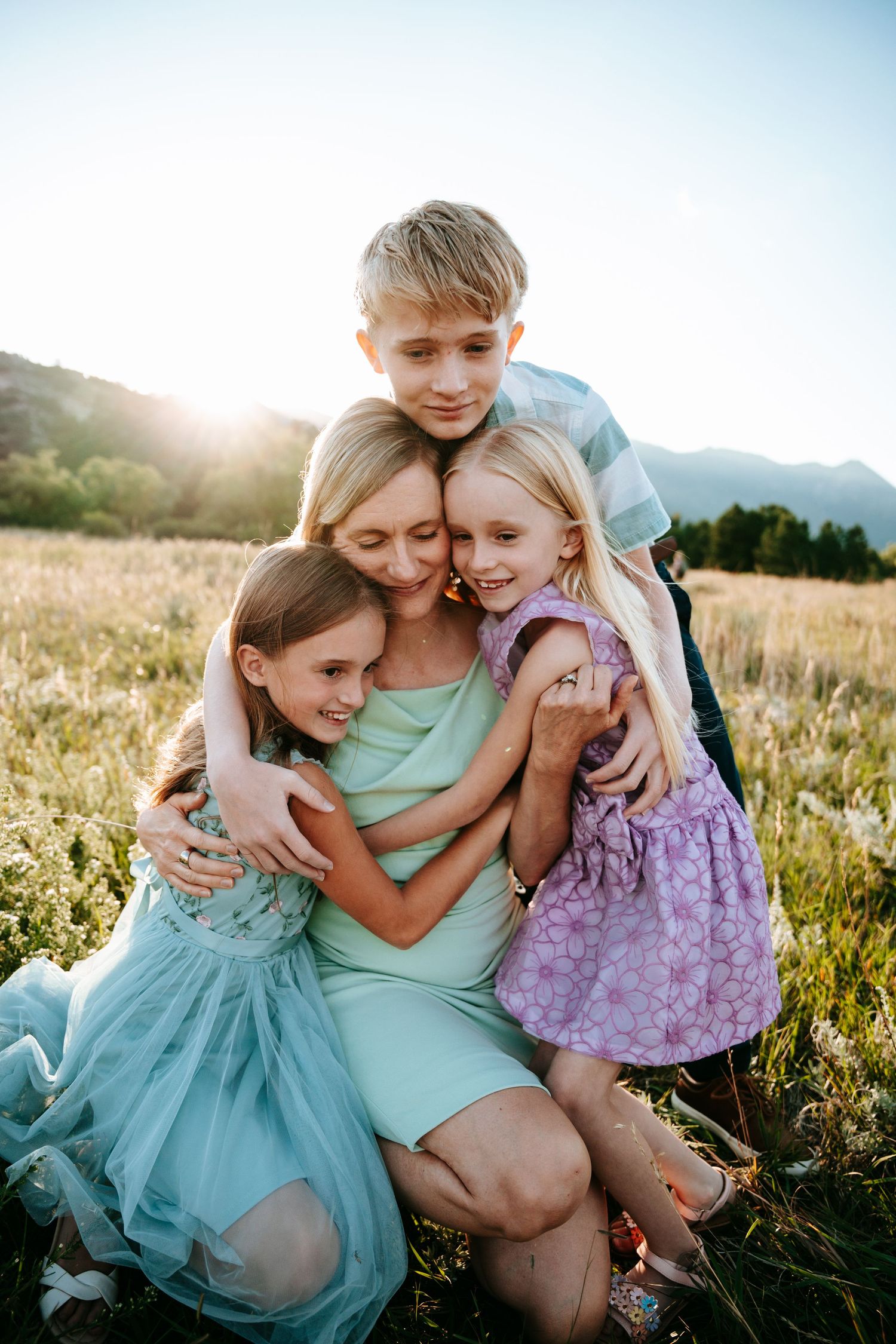 Blonde mother kneeling in sunlit field embracing three children in a warm family group hug.