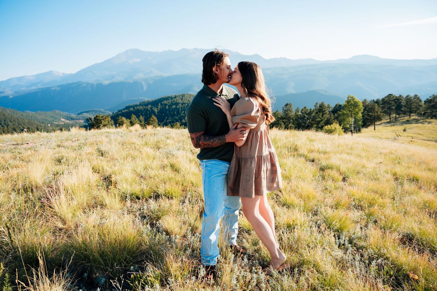 Couple kissing in a golden meadow with Rocky Mountain peaks in the background during a sunny engagement session.