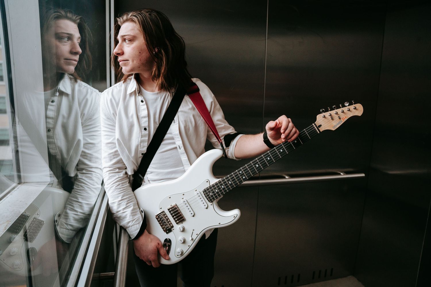 Young man with long hair holding a white electric guitar inside a modern elevator.