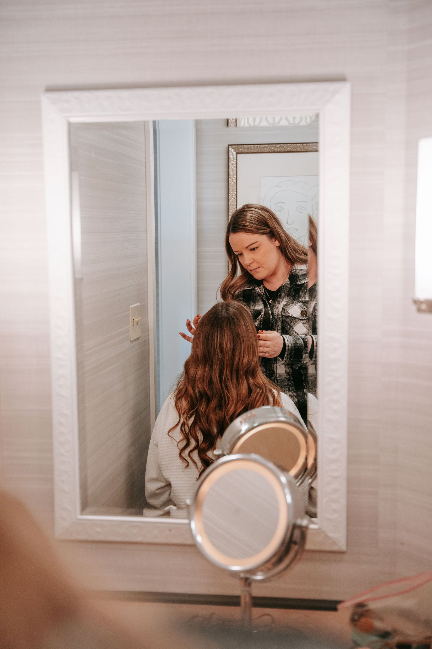 Hairstylist styling a bride's long wavy brown hair in a bathroom mirror reflection.