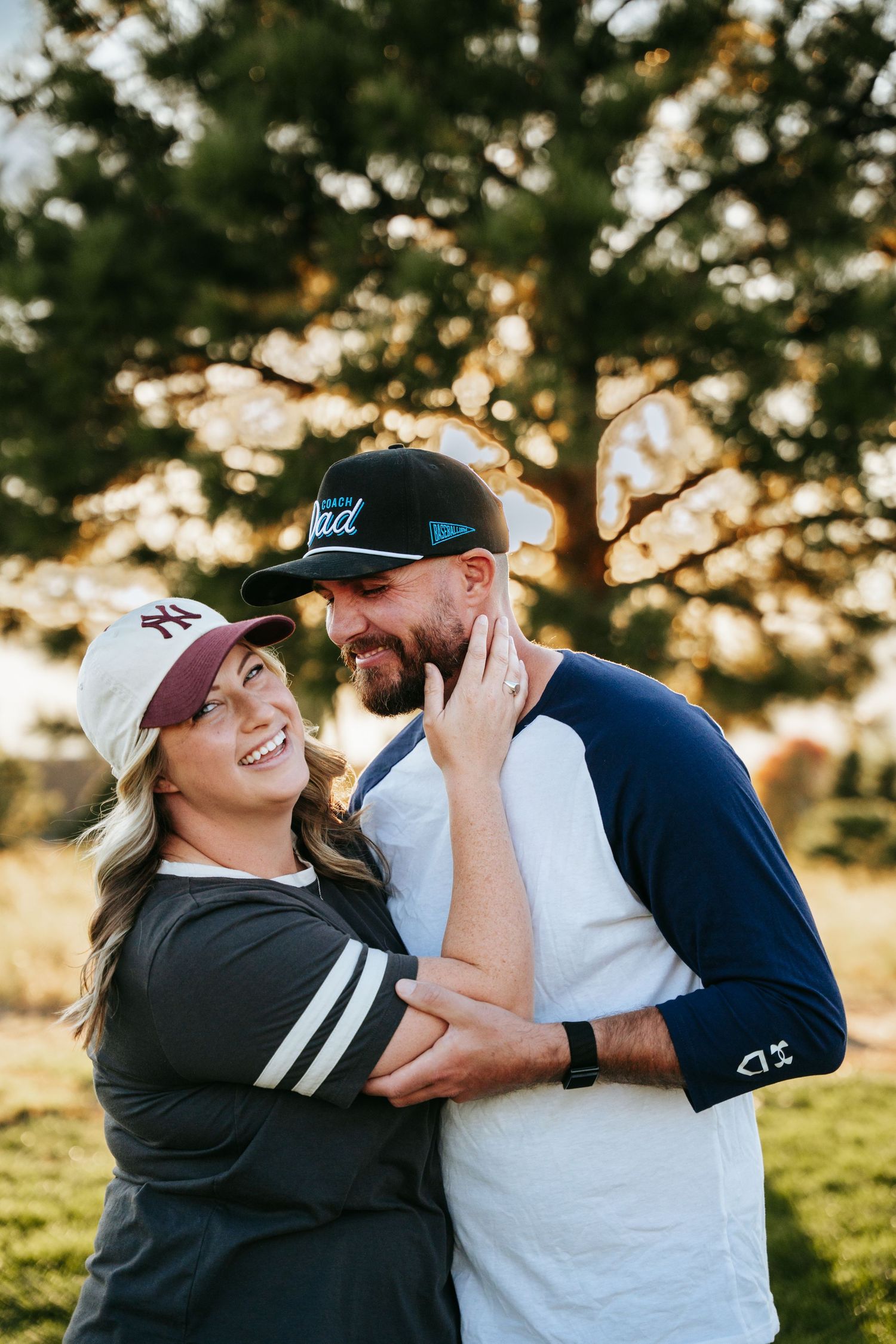Smiling couple in baseball caps and raglan tees embracing outdoors in golden sunlight with evergreen trees and a grassy field behind them.