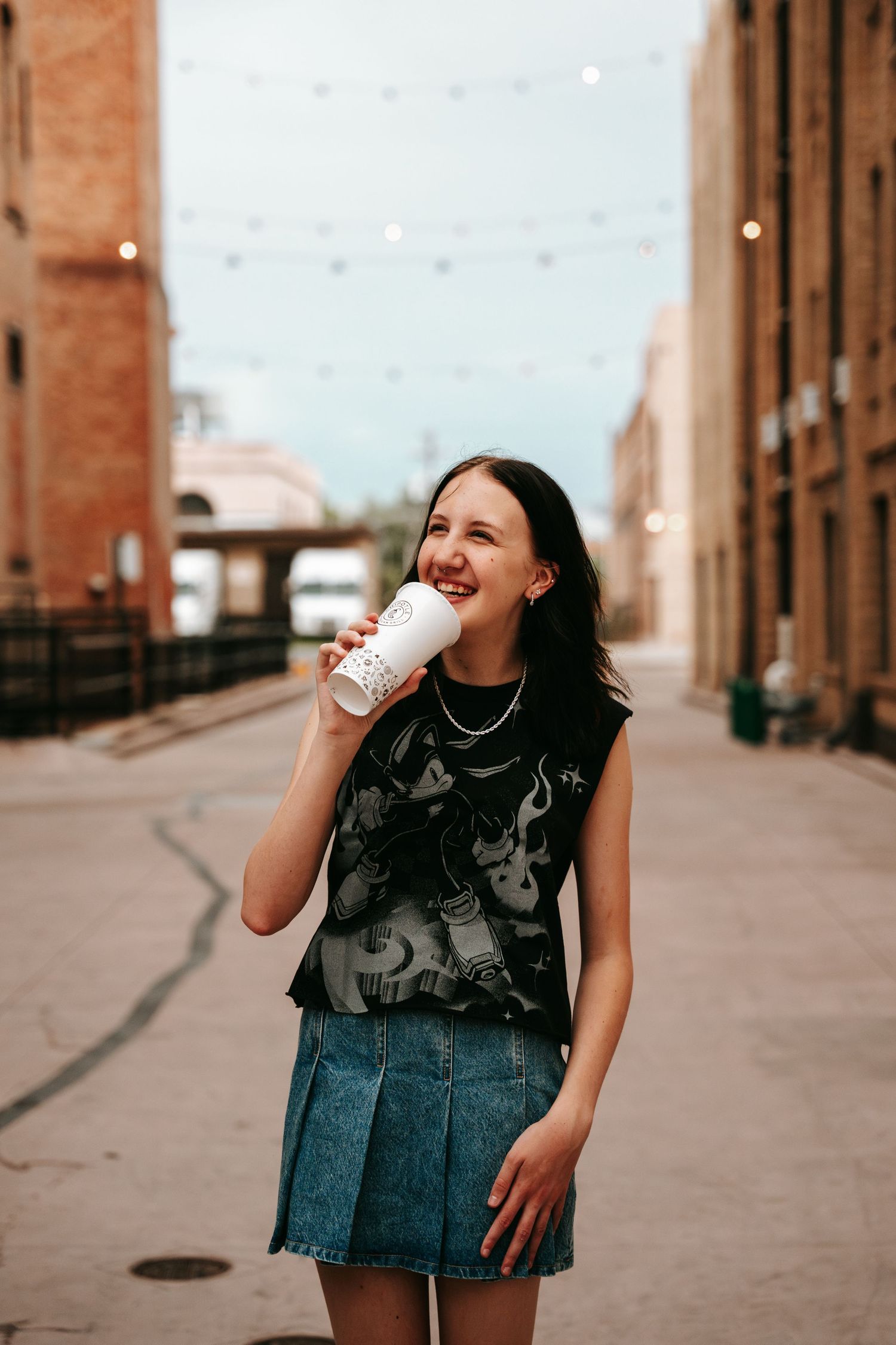 Laughing young woman holding coffee cup in urban alley with string lights.