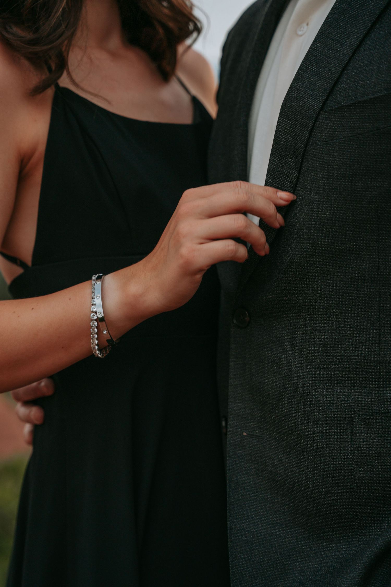 A woman in a black dress with a silver bracelet gently touches a man's dark gray suit jacket.