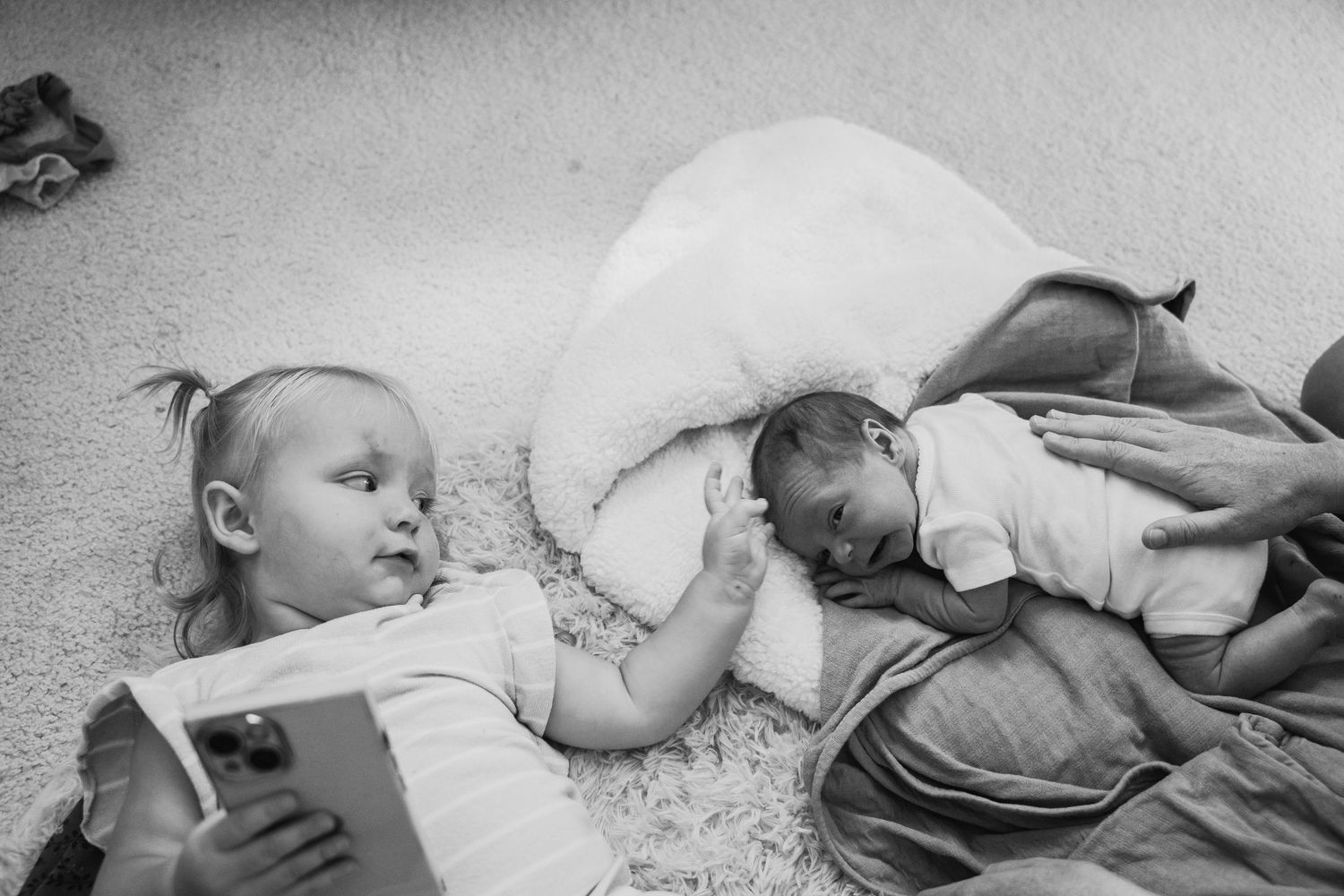 Black and white photo of toddler reading beside sleeping newborn baby on carpet floor.