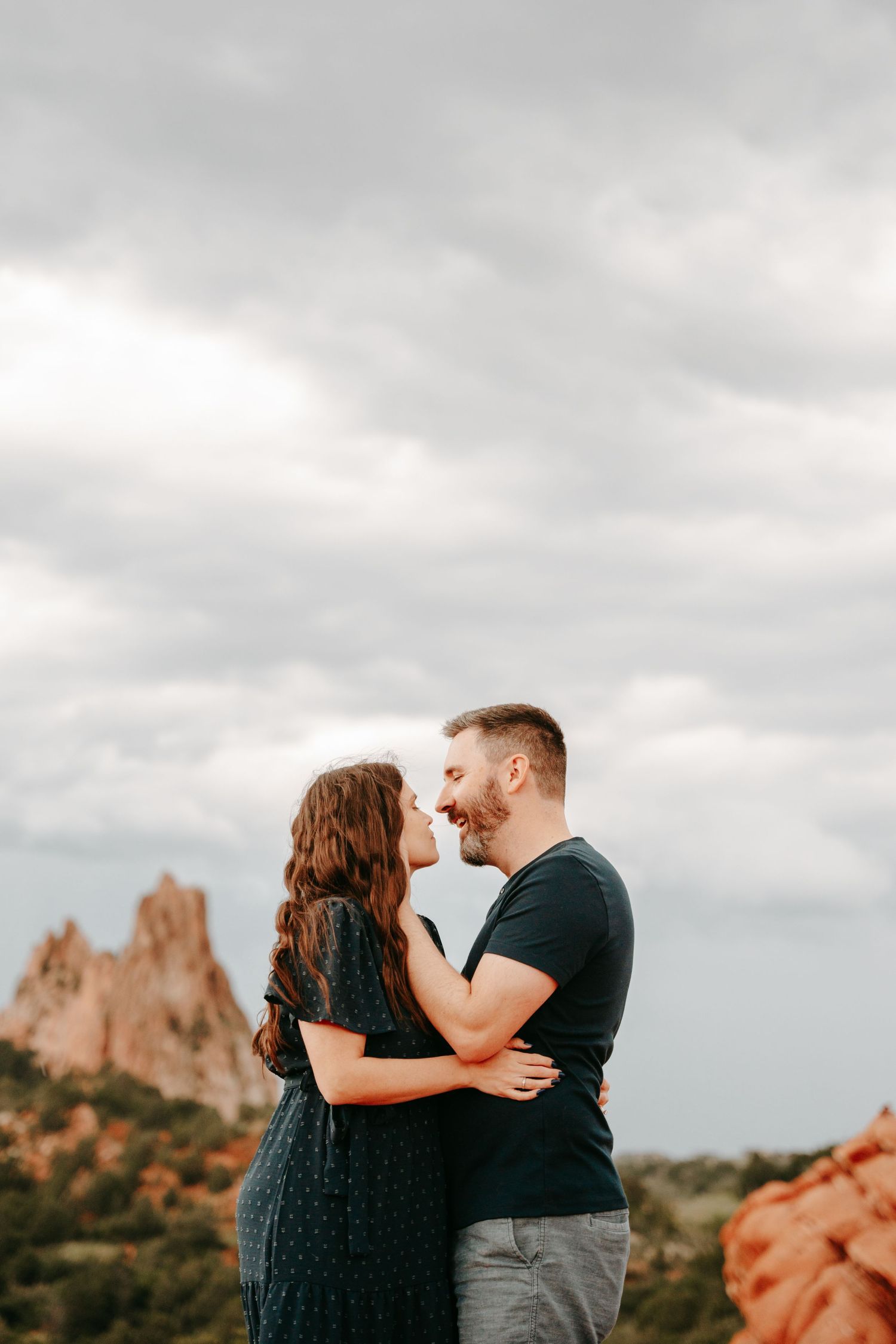 Couple embracing and smiling at each other with red rock formations under cloudy sky.