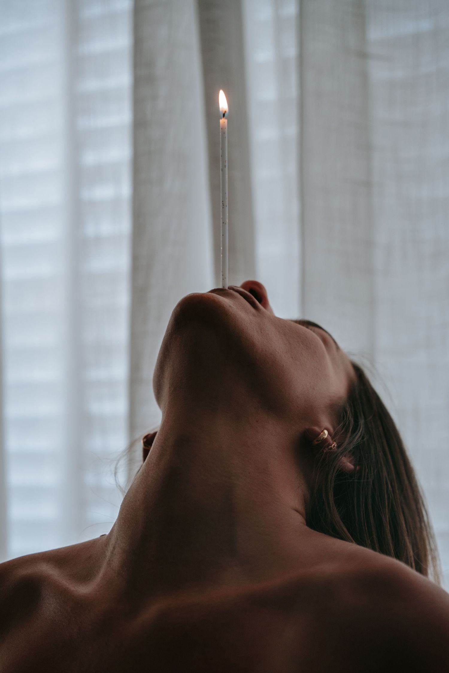 Woman balancing a lit candle while looking upward against sheer white curtains.