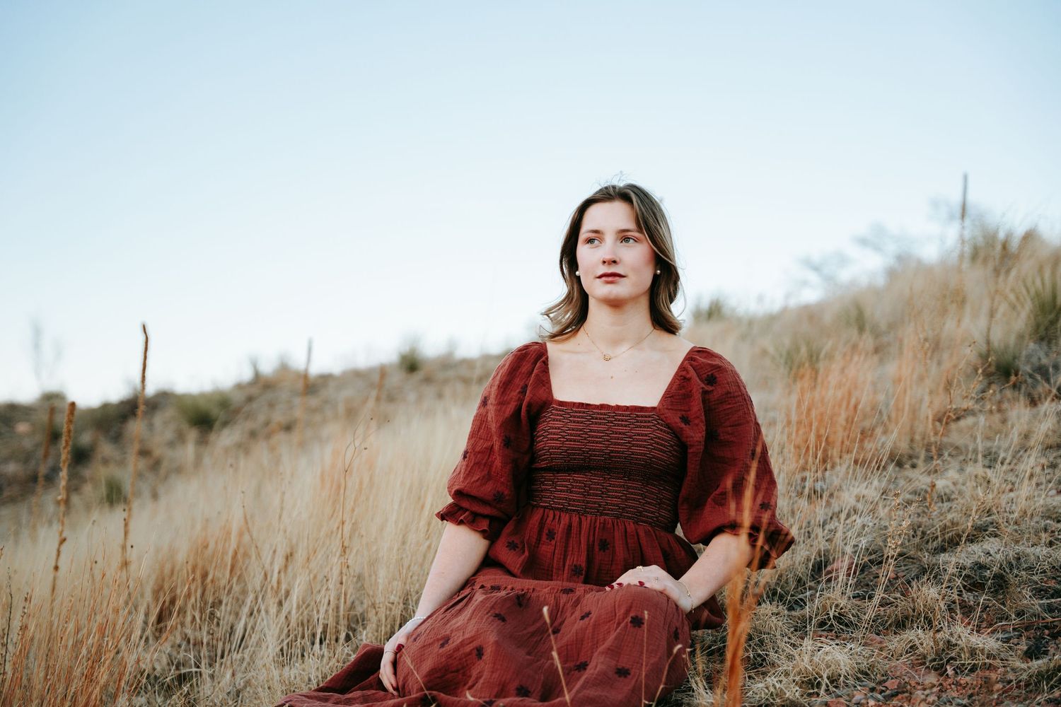 Young woman in red smocked dress sitting in field of dry grass under clear sky.