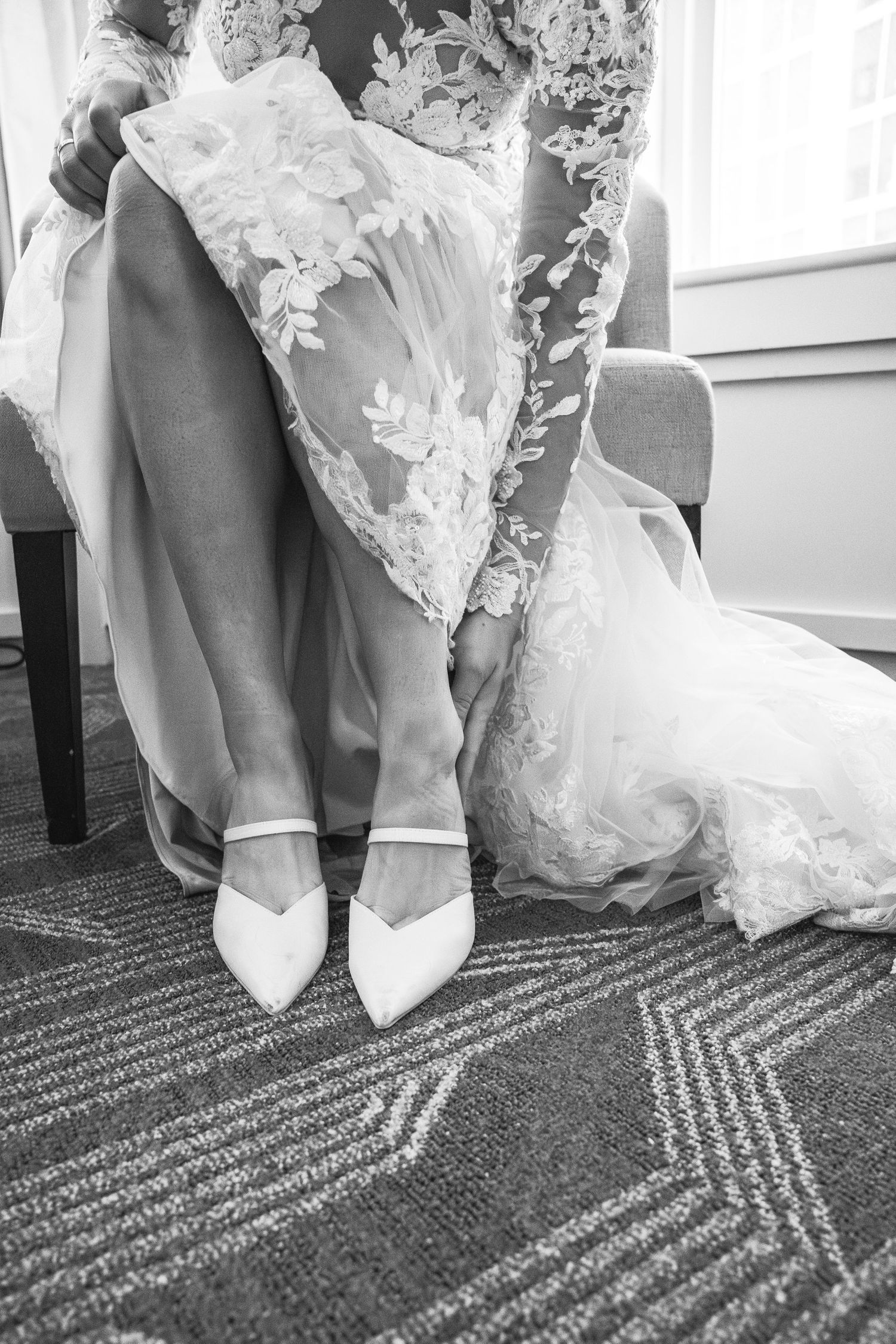 Black and white photo of bride in lace gown putting on white pointed-toe heels.