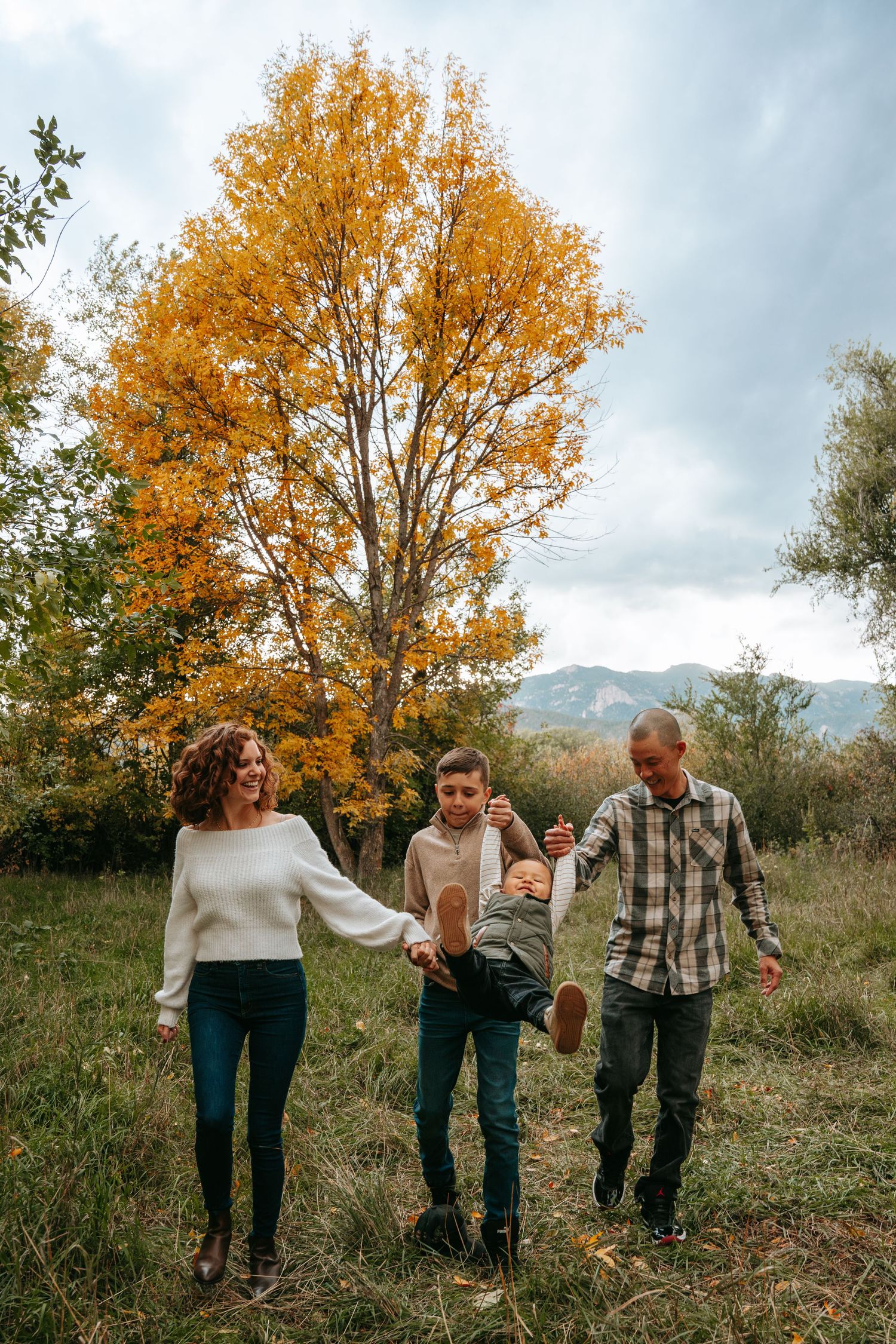 Family of four swinging toddler outdoors in autumn park with golden trees and mountain backdrop.