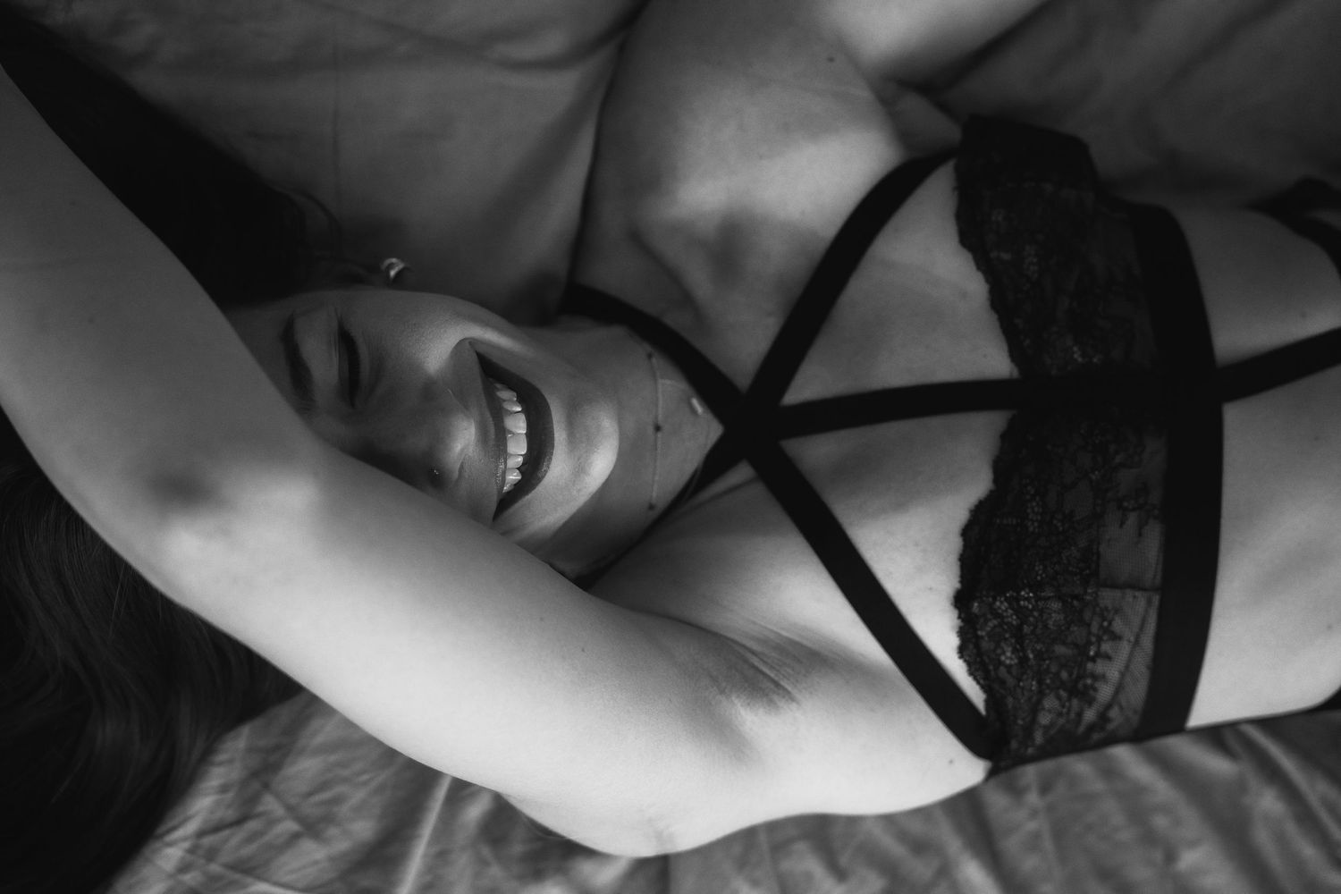 Black and white boudoir photo of a smiling woman lying on a bed wearing lace lingerie.